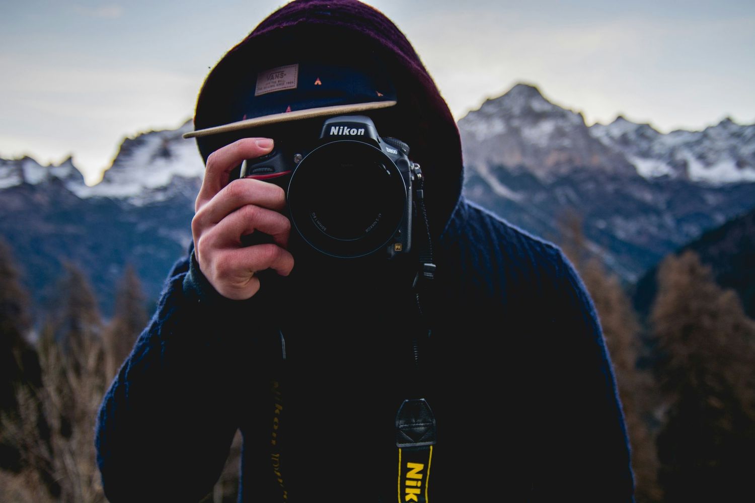 a man in a hoodie and baseball cap standing in from of a mountain range hides behind his Nikon camera