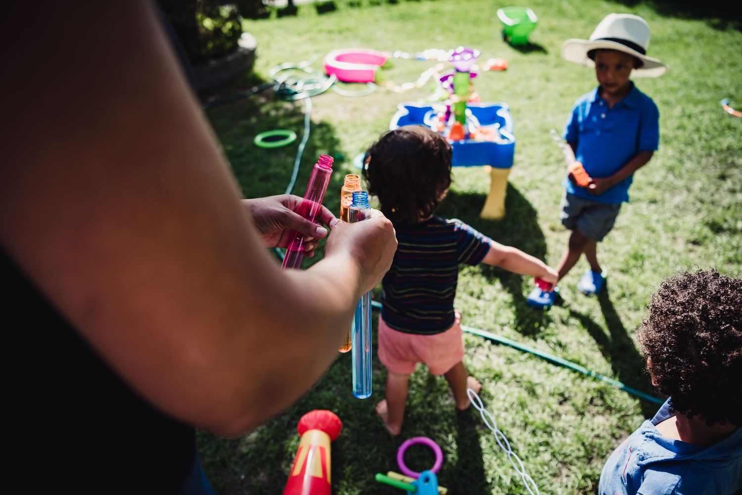 Colour photo of a woman holding 3 bubble bottles as kids play in the background on green grass.