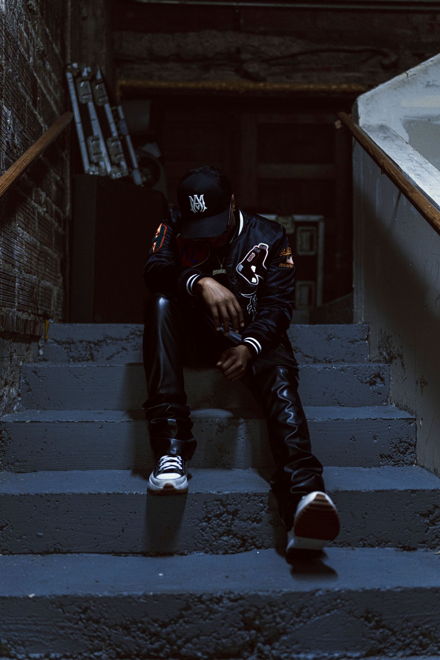 Moody urban portrait on concrete steps with varsity jacket and baseball cap in dark lighting.