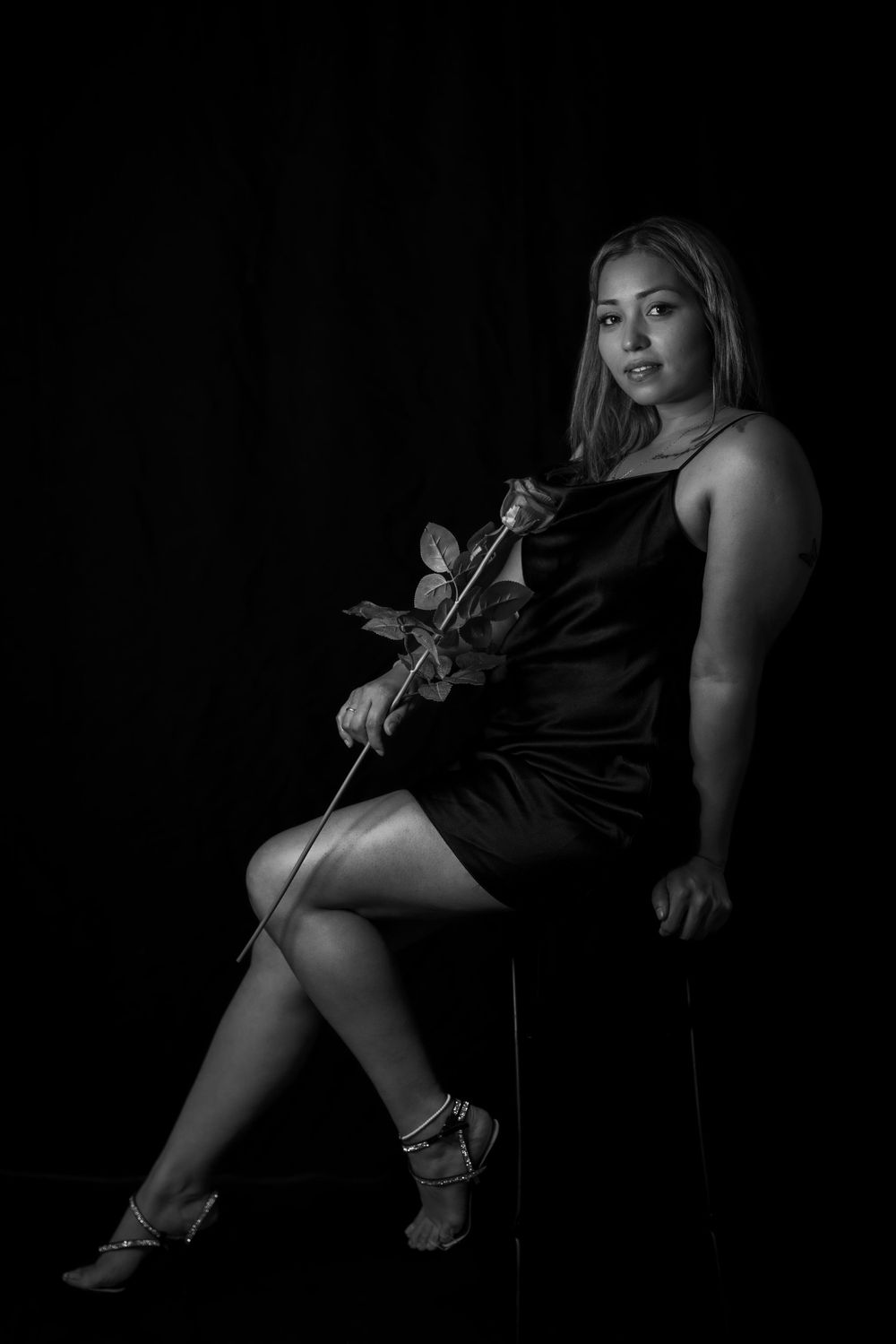 Moody portrait of woman in black dress holding rose against dark backdrop.