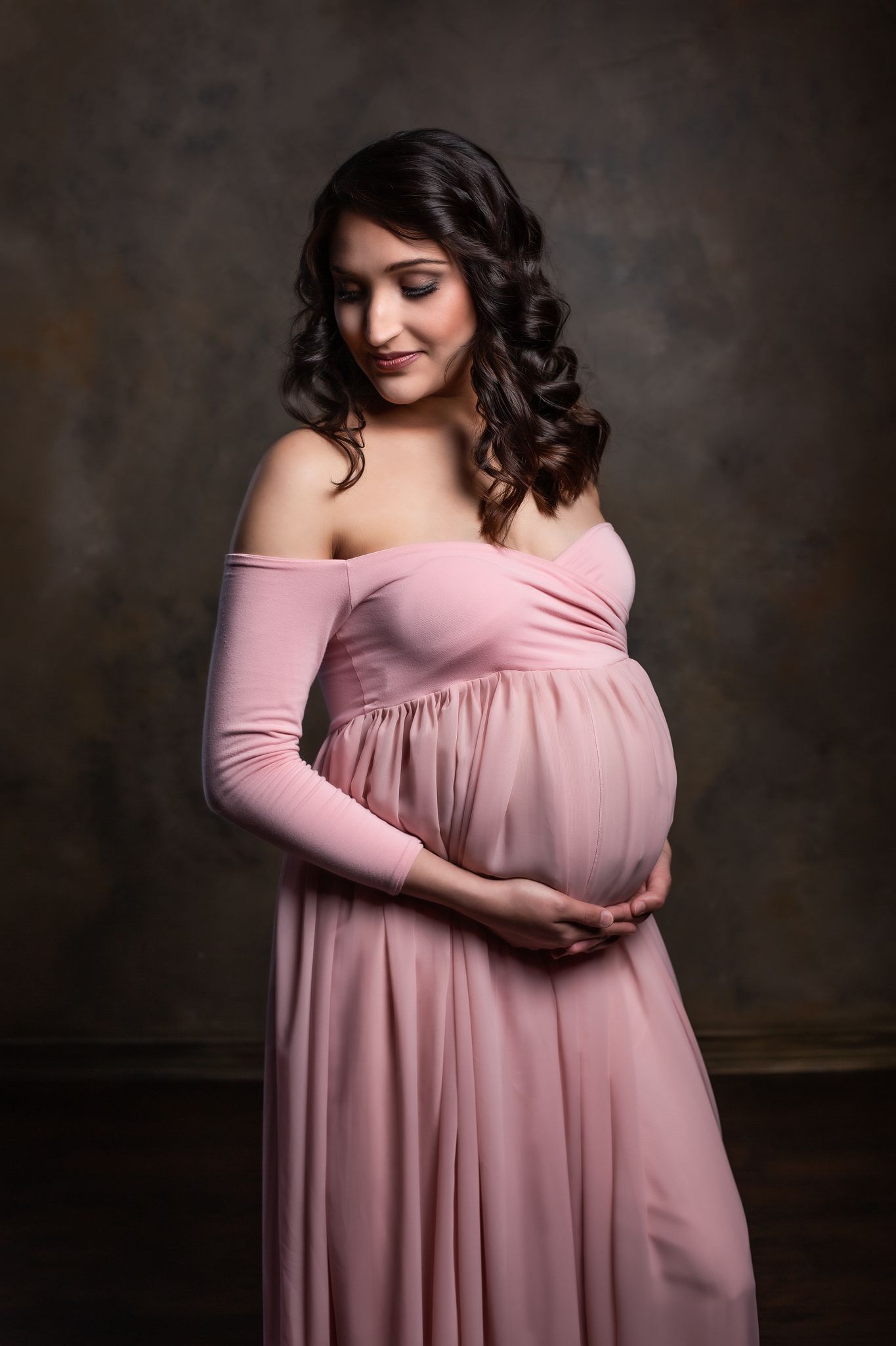 Elegant maternity portrait in pink off-shoulder gown against dark backdrop.