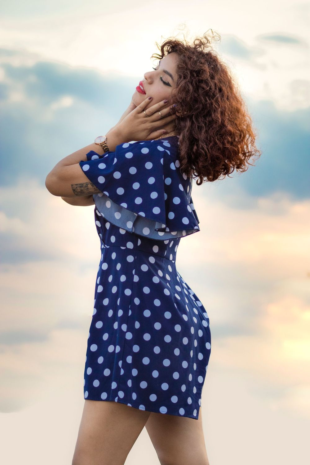 Woman in blue polka dot dress posing against a dramatic sunset sky with curly hair and red lips.