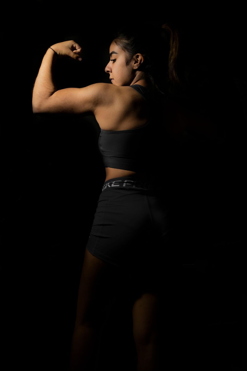 Athletic woman in black sportswear flexing bicep muscle against dark studio background.