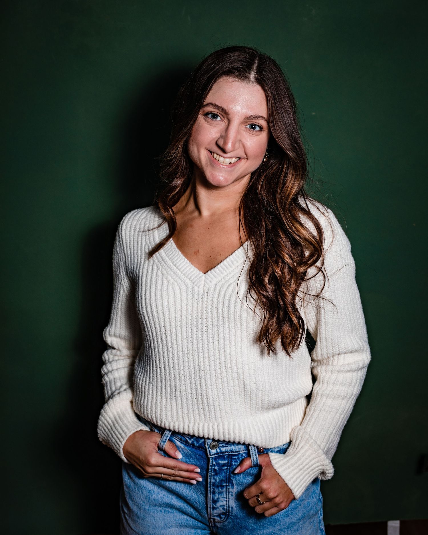 Professional portrait of someone in a white sweater and jeans against a dark green background.