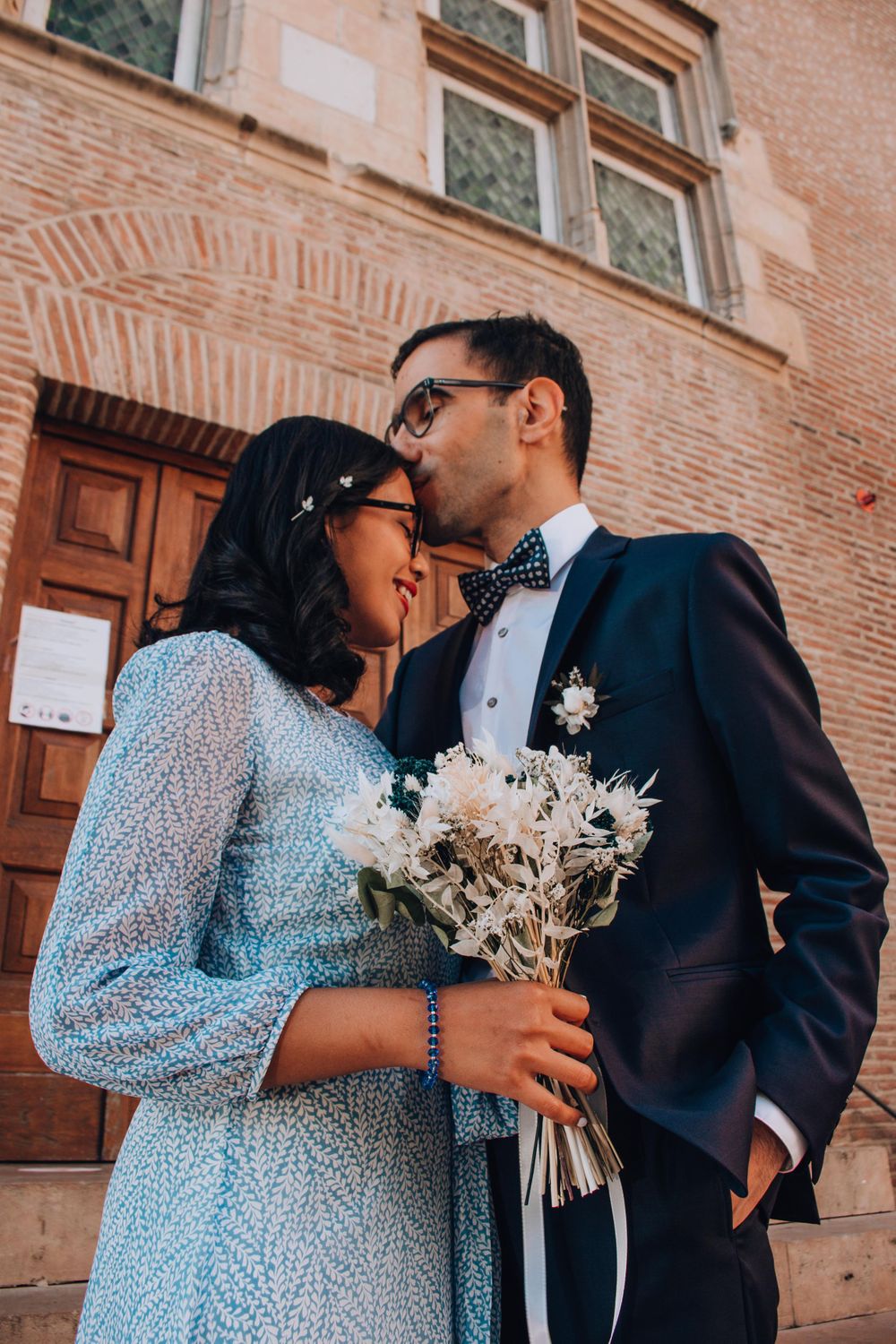 A couple shares a tender moment while holding a white floral bouquet against a brick building.