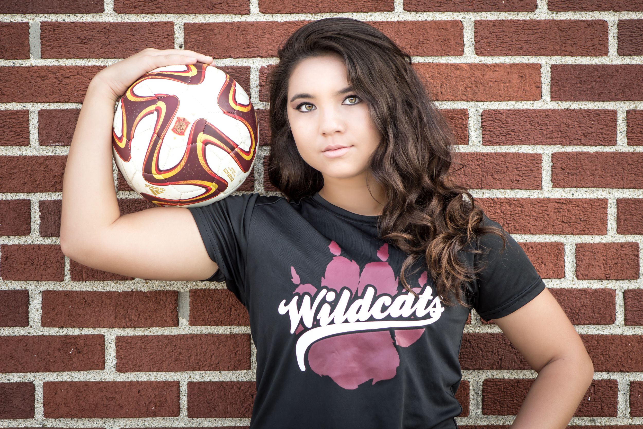 Teen girl in Wildcats shirt holding soccer ball against brick wall for portrait.