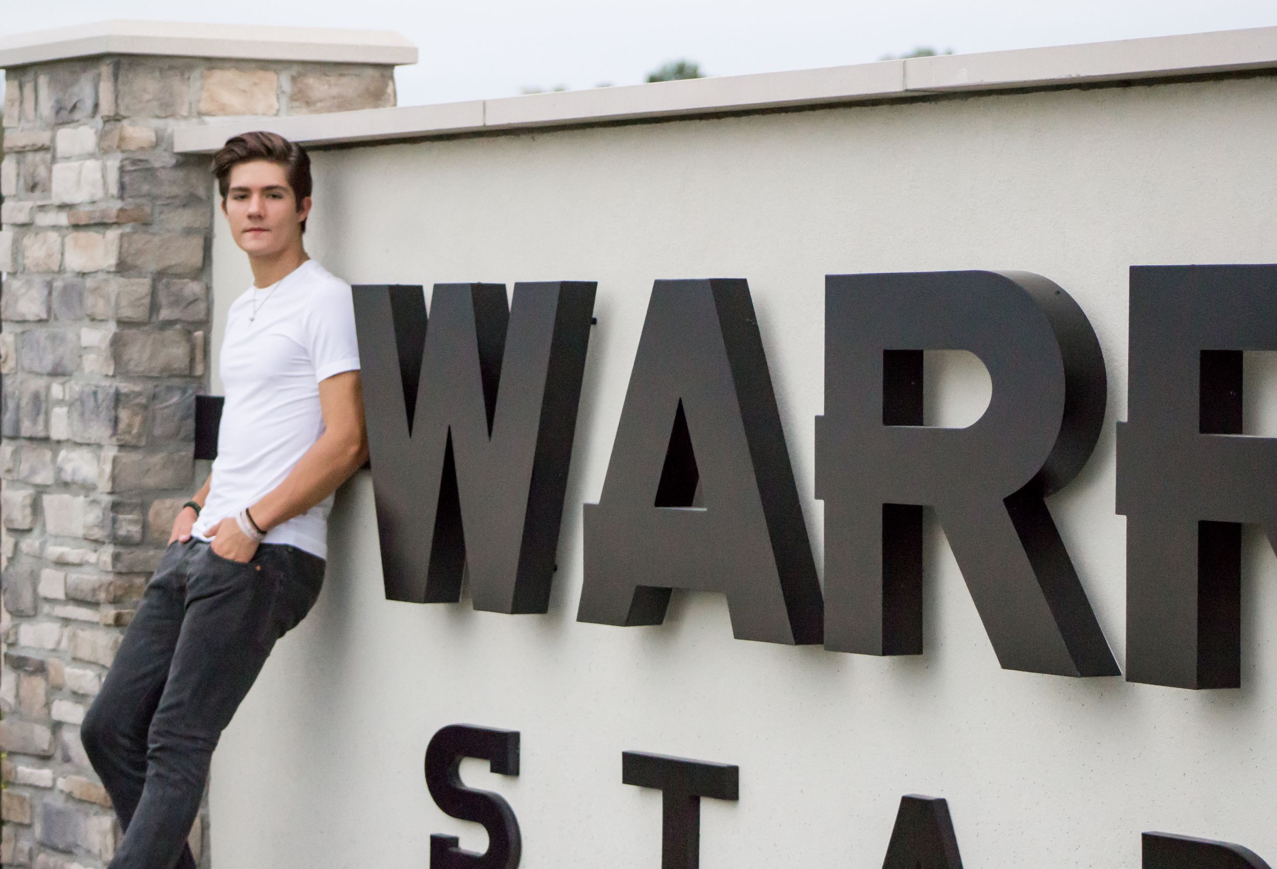 Teenage boy in white t-shirt leans against a Warriors Stadium entrance sign outdoors.