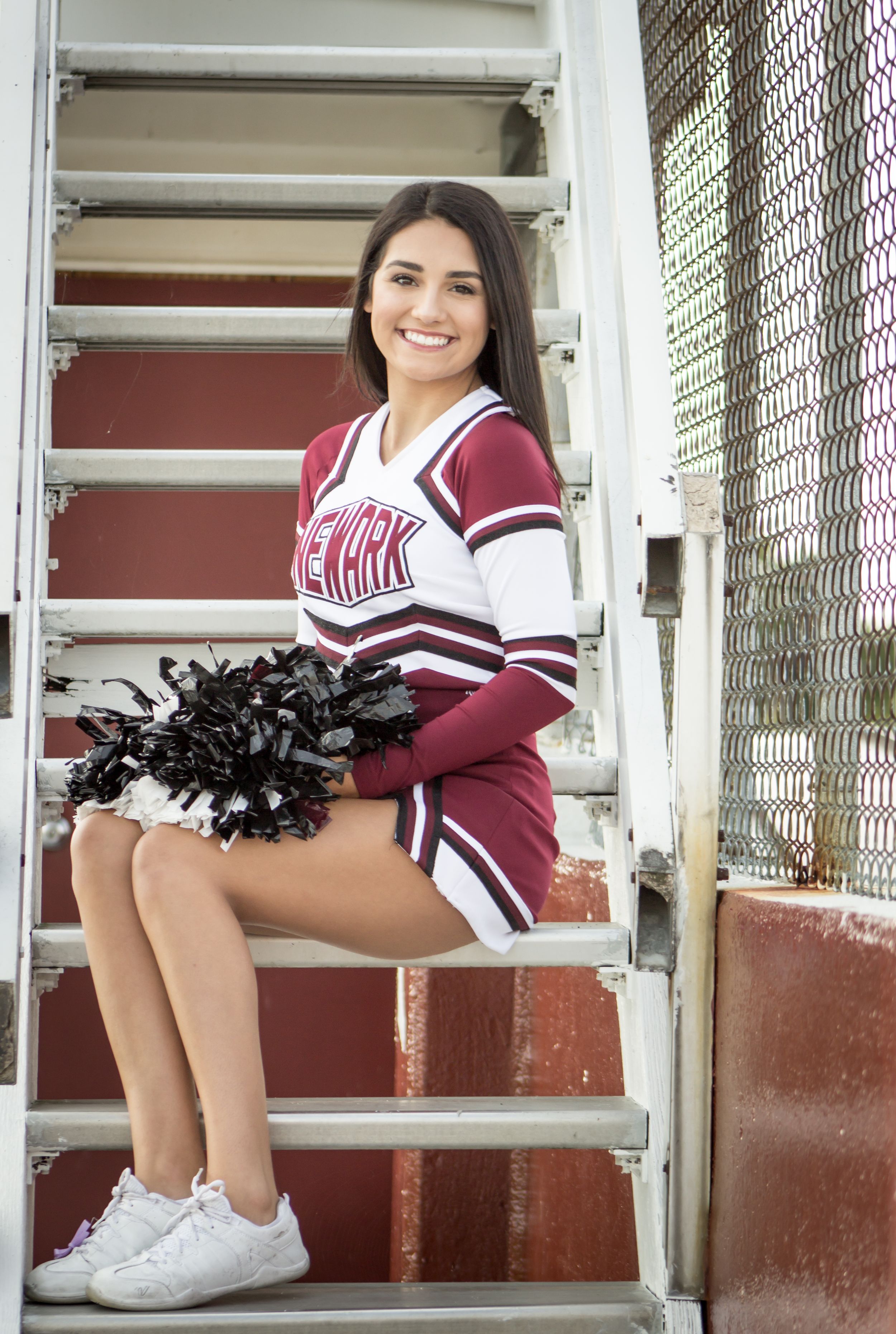 Newark cheerleader sitting on stadium stairs holding black pom-poms, smiling brightly.