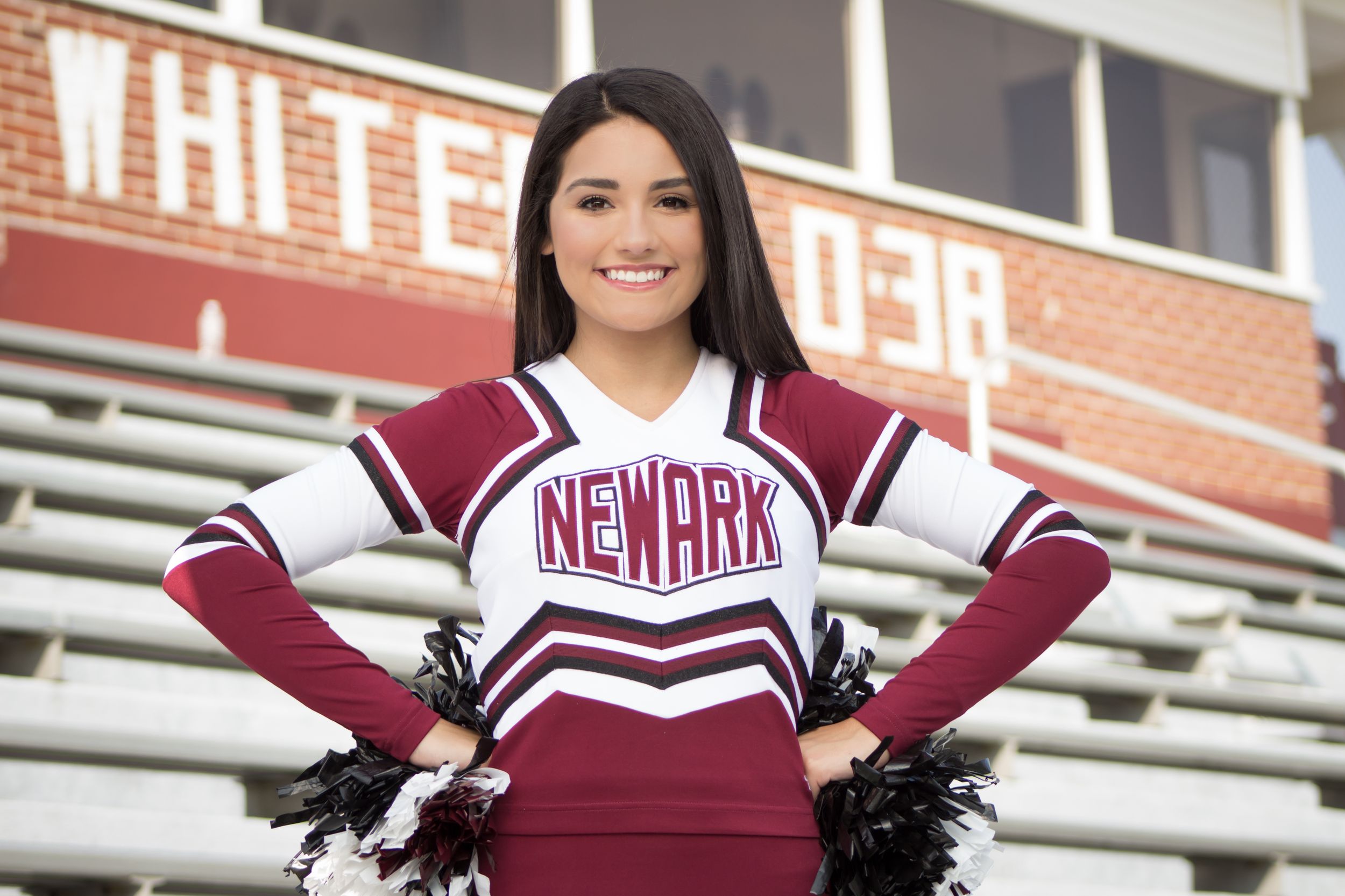 Newark cheerleader in maroon and white uniform poses with pom-poms at a football stadium.
