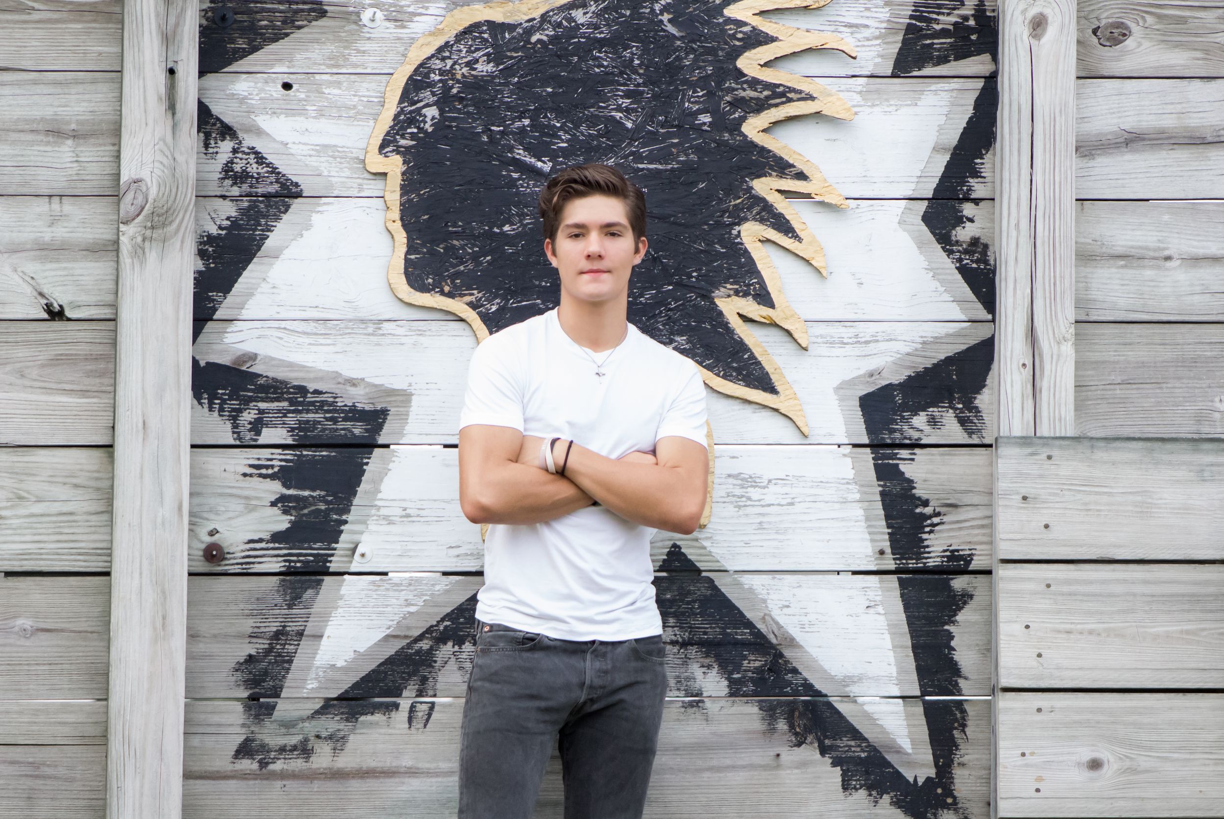Young man in white t-shirt poses with arms crossed before a painted warrior school mascot mural.