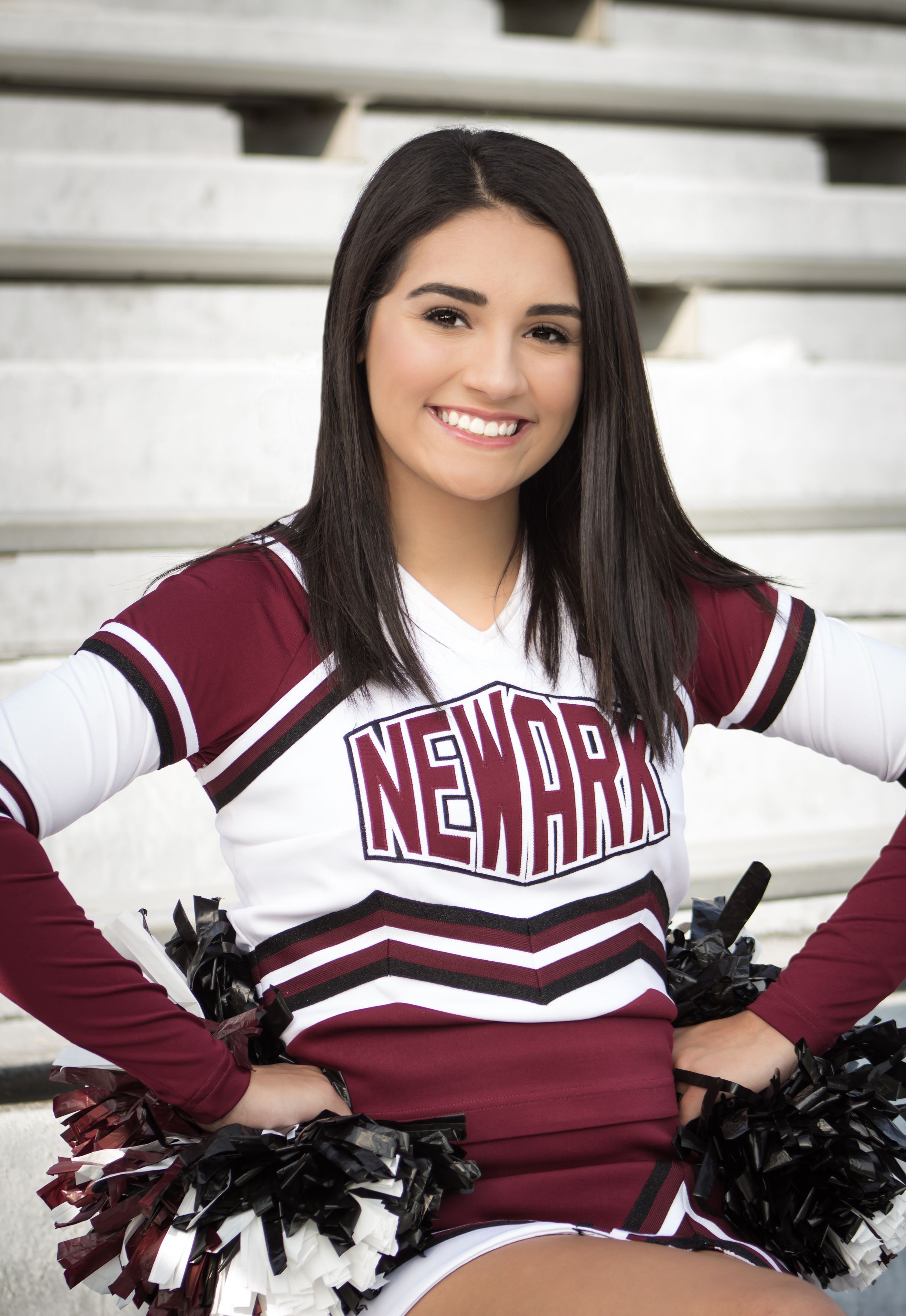 Smiling Newark cheerleader in maroon uniform holds pom-poms while seated in stadium bleachers.