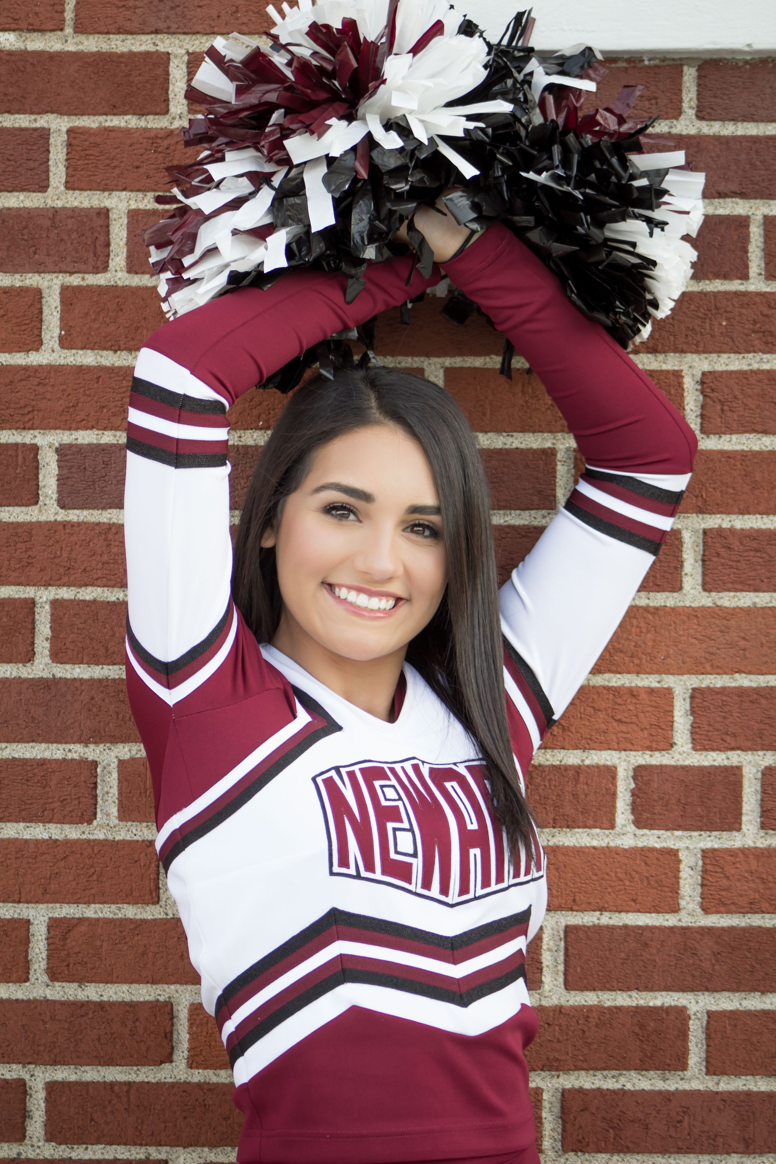 Newark cheerleader raises maroon and white pom-poms overhead against a brick wall backdrop.