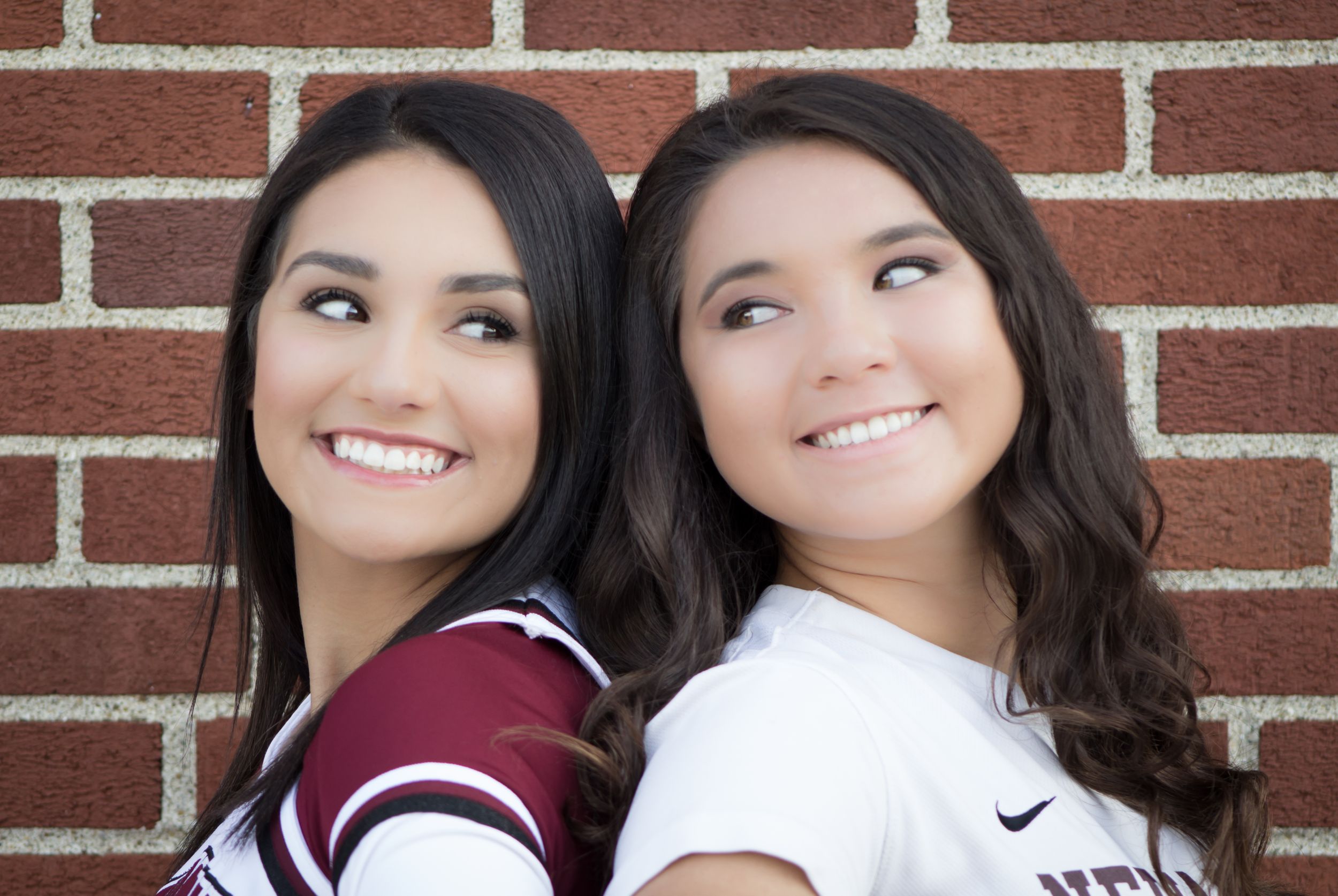 Two smiling young women in Newark sports uniforms pose back-to-back against a brick wall.