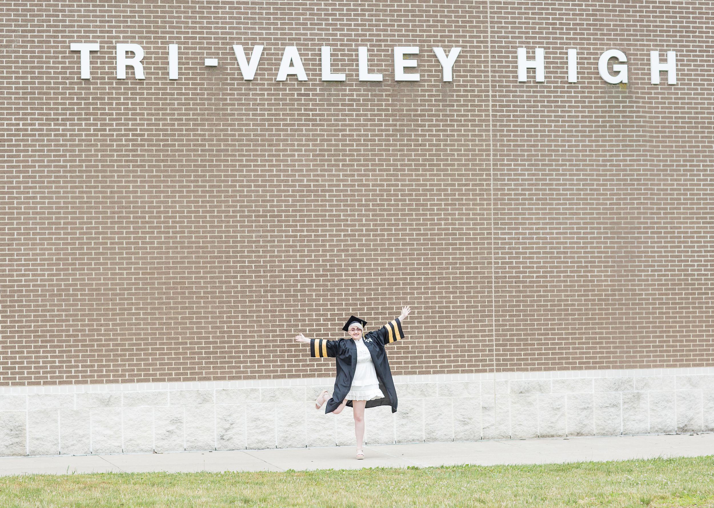 Graduate in cap and gown jumps joyfully in front of Tri-Valley High School brick building.