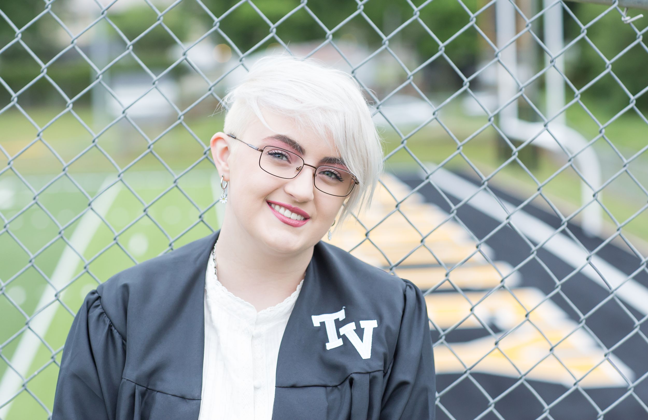 Smiling graduate in black cap and gown posing by a chain-link fence outdoors.
