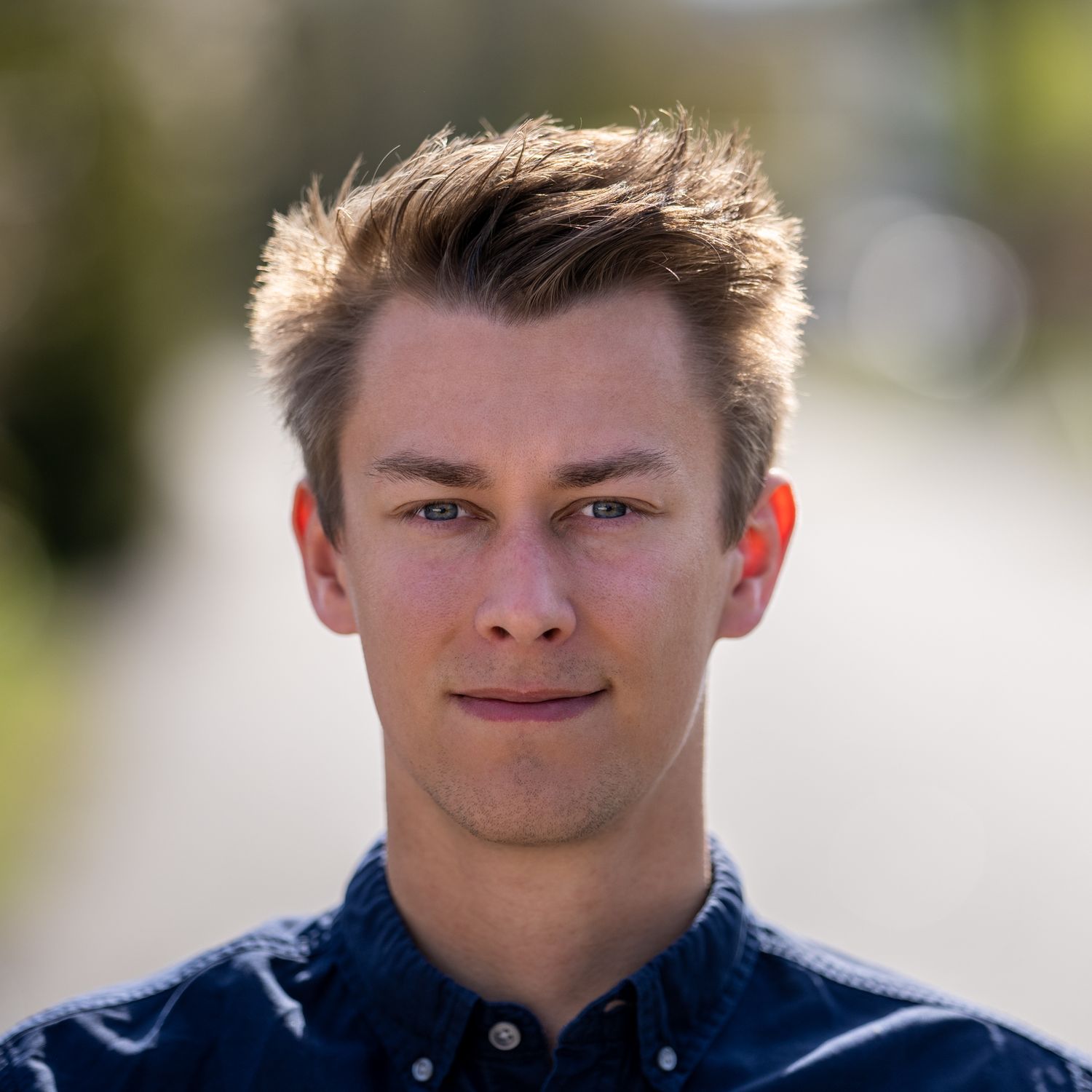 Professional headshot portrait of a young person in a navy blue shirt against a blurred outdoor background.
