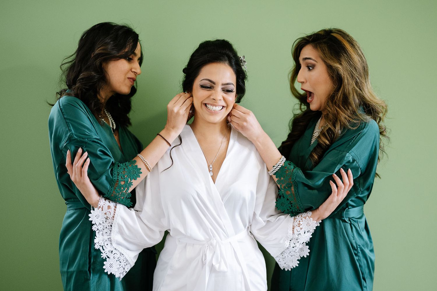 Three women in teal and white robes sharing a joyful moment against a mint green wall.