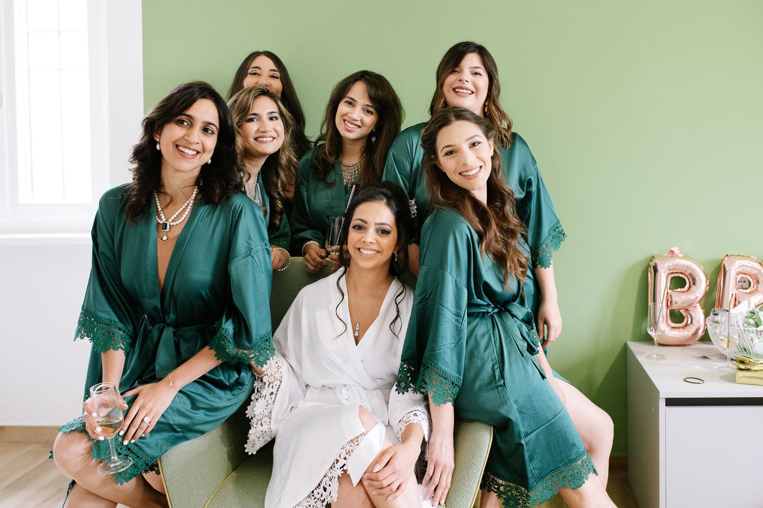 Group wearing coordinated teal robes and one white robe gather for pre-wedding preparation photo.