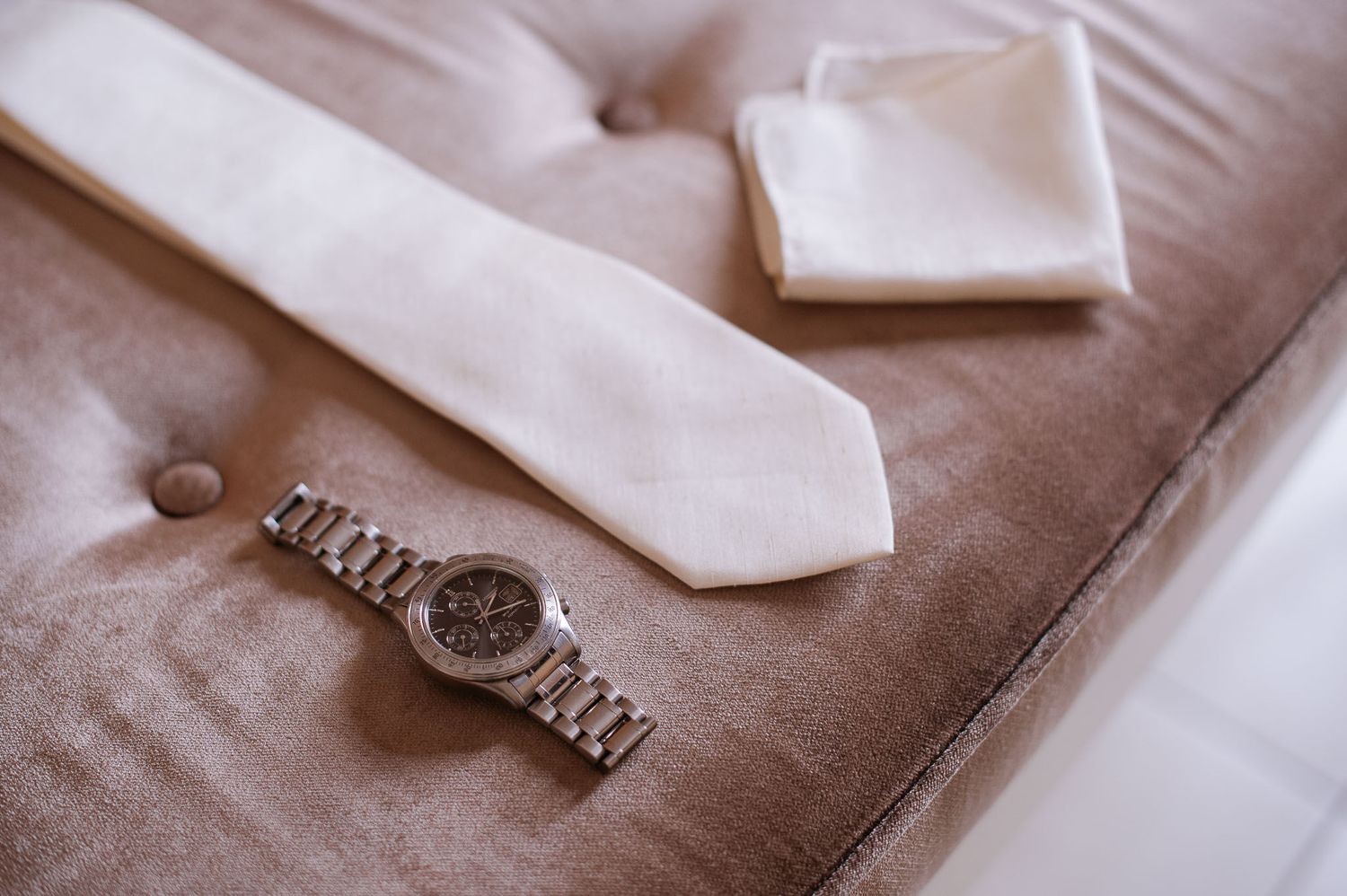 Silver watch and white tie arranged neatly on brown velvet surface.