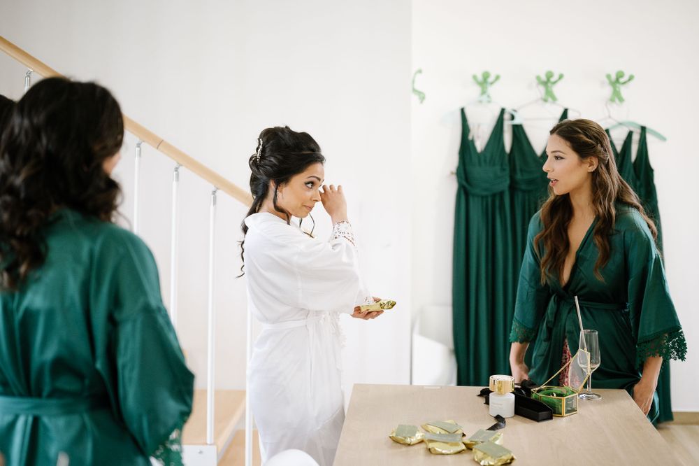 Women in teal robes and white robe prepare for special event with emerald dresses hanging in background.