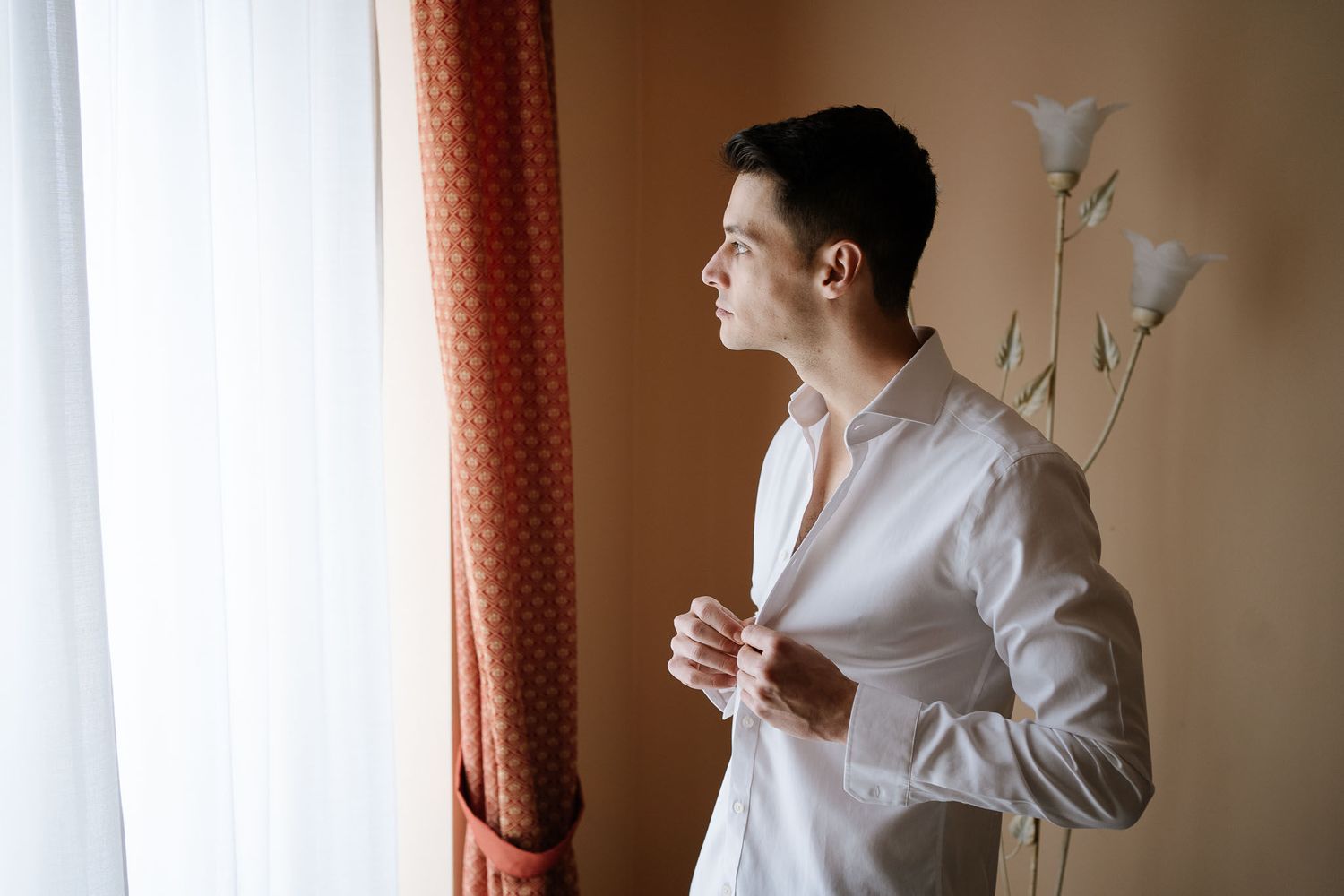 Person in white dress shirt adjusts collar while standing by window with orange curtains.