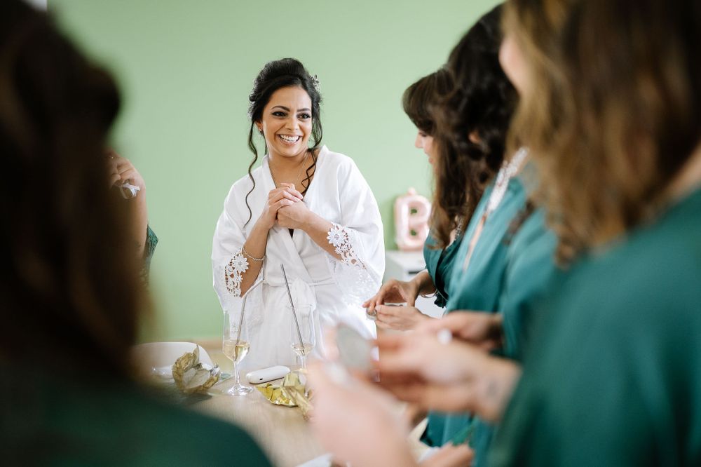Woman in white robe clasps hands joyfully while surrounded by bridesmaids in green robes.