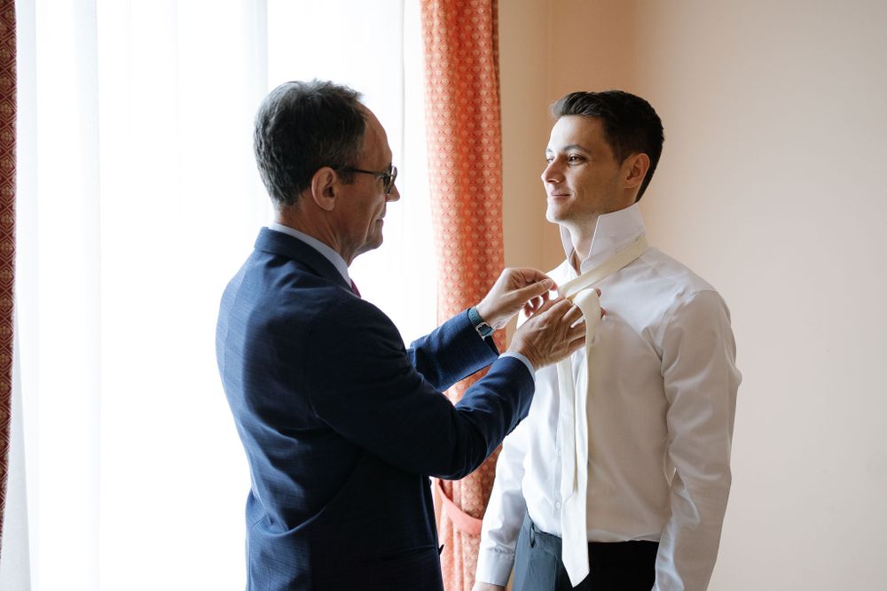 Older man helps adjust younger man's tie during formal dressing preparation.