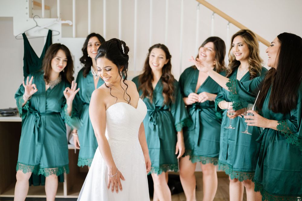Bride in white gown poses while bridesmaids in teal robes laugh in background.