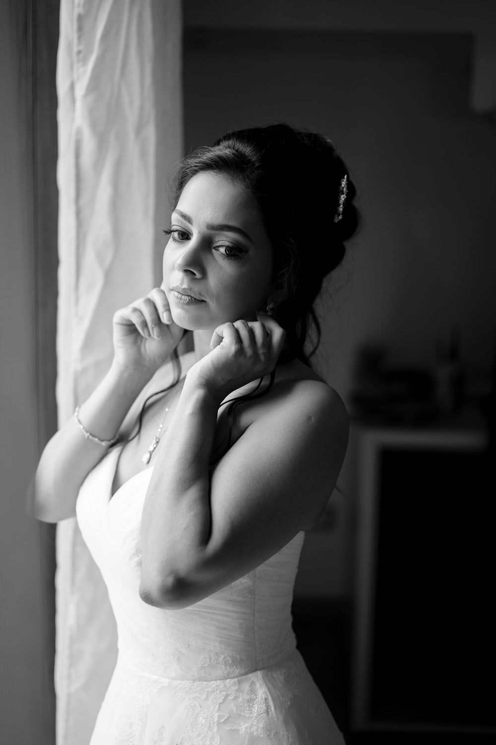 Black and white portrait of bride in wedding dress adjusting jewelry by window.