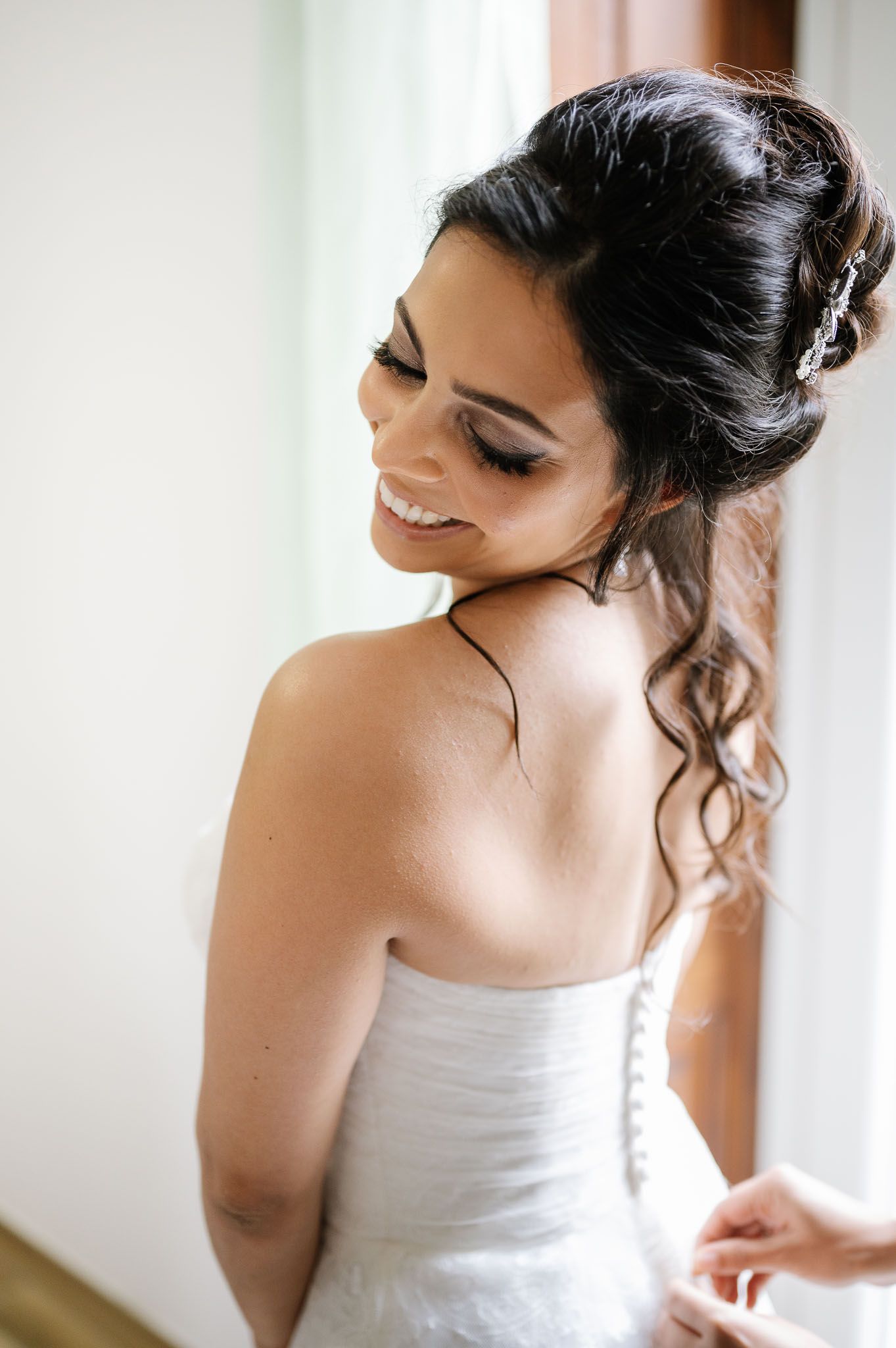 A bride in a white dress smiles while looking down, showing her elegant updo hairstyle with loose curls.