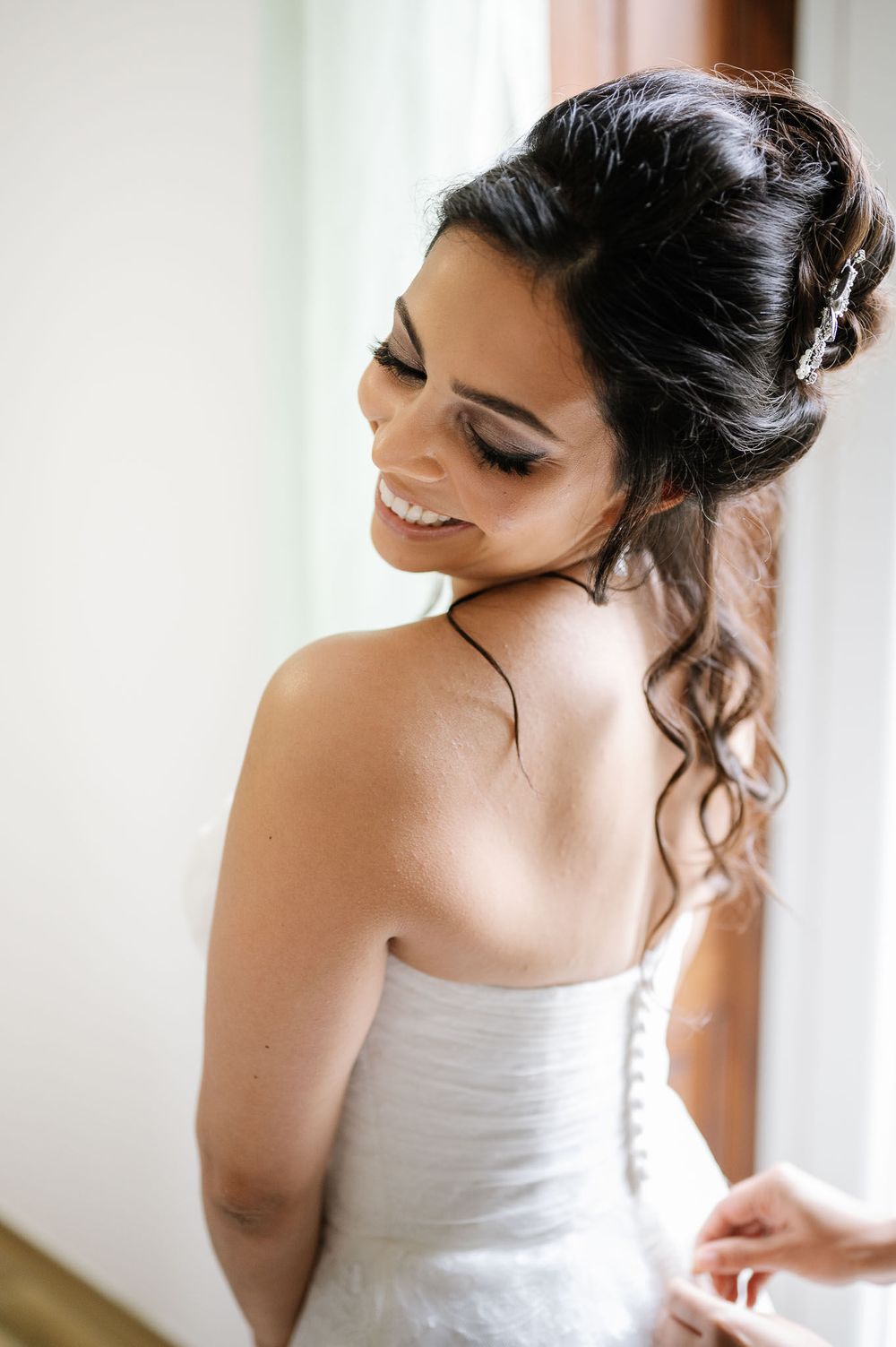 A bride in a white dress smiles while looking down, showing her elegant updo hairstyle with loose curls.