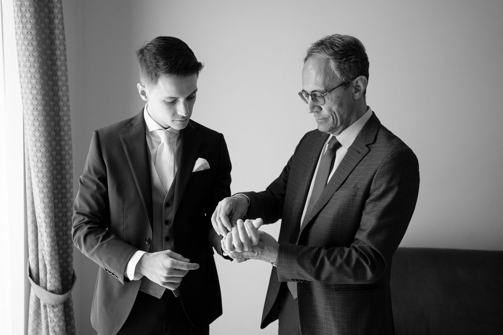 Black and white photo of two men examining wristwatch during wedding preparations.