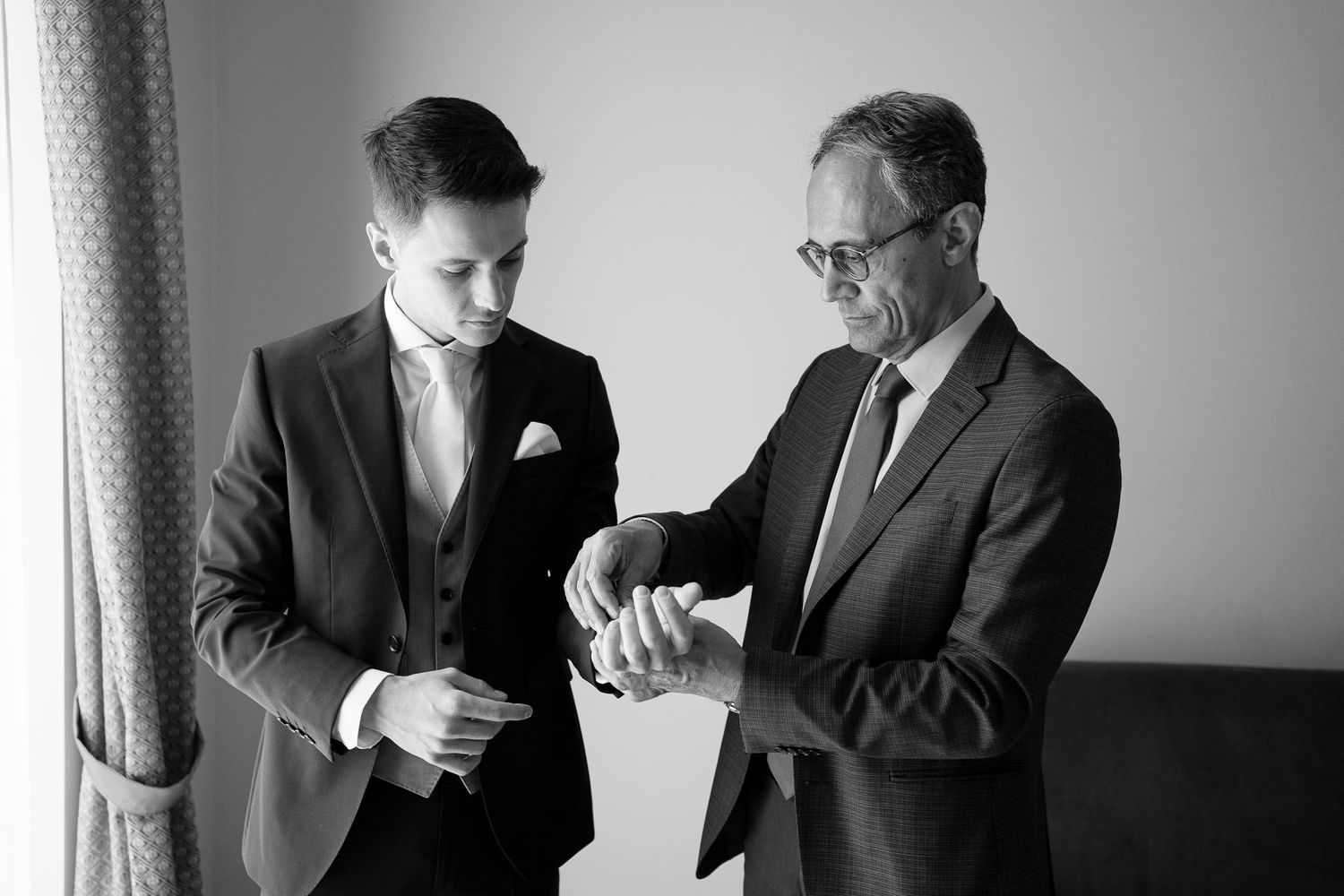 Black and white photo of two men examining wristwatch during wedding preparations.