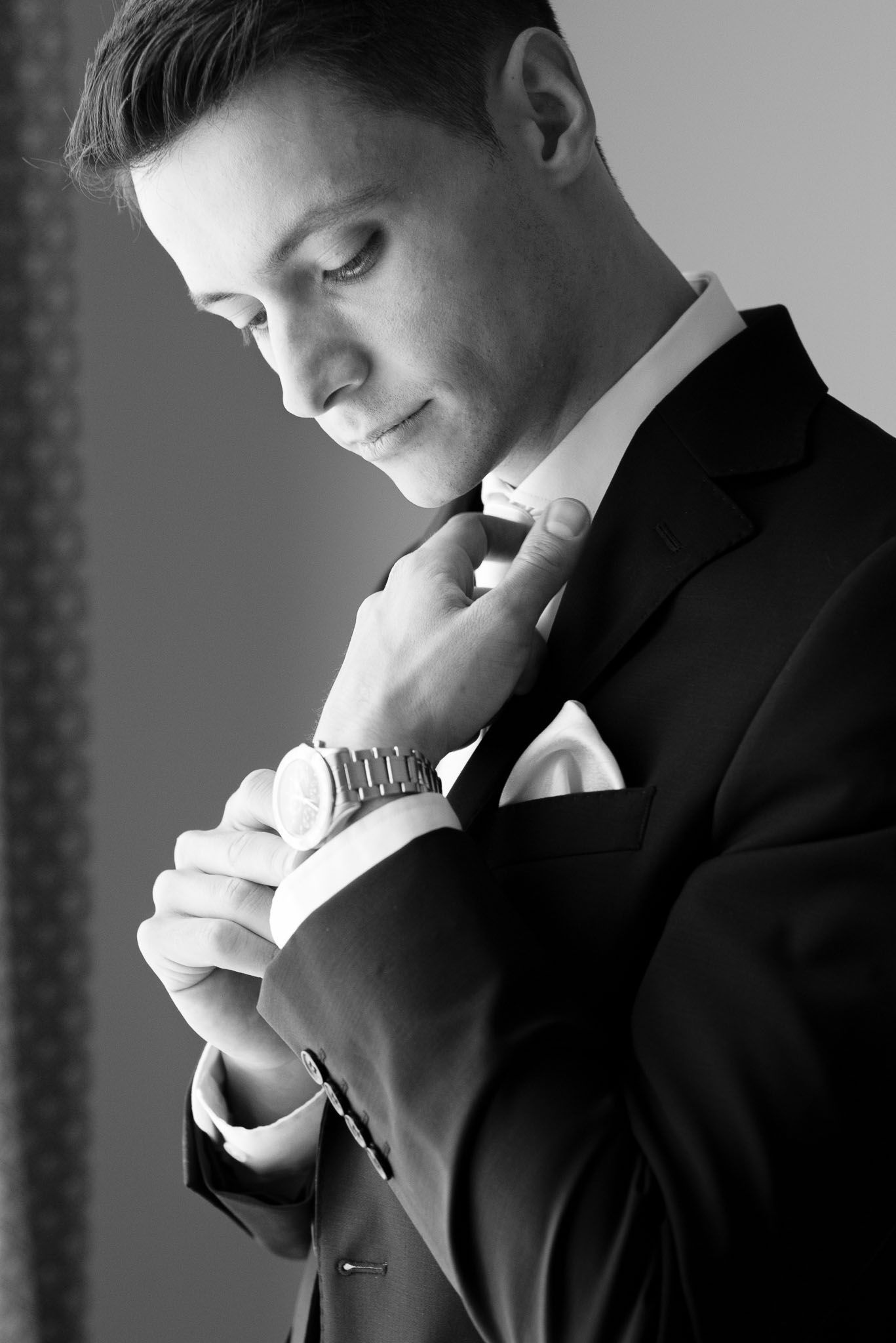 A groom in a black tuxedo adjusts his tie in a reflective black and white portrait.