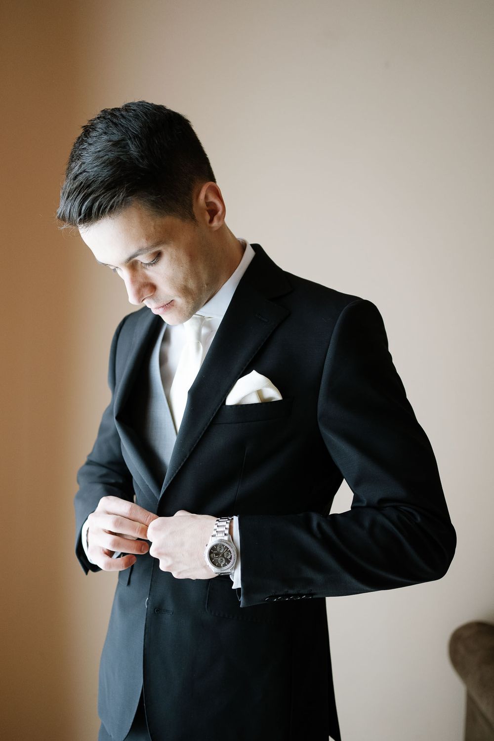 Groom in a classic black suit adjusts his cufflinks while standing in natural light.