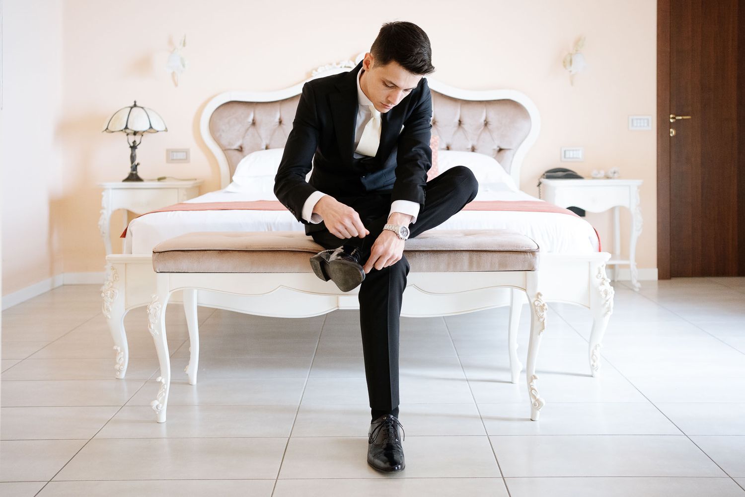 Groom in formal attire sits on an elegant white sofa while adjusting his shoe.