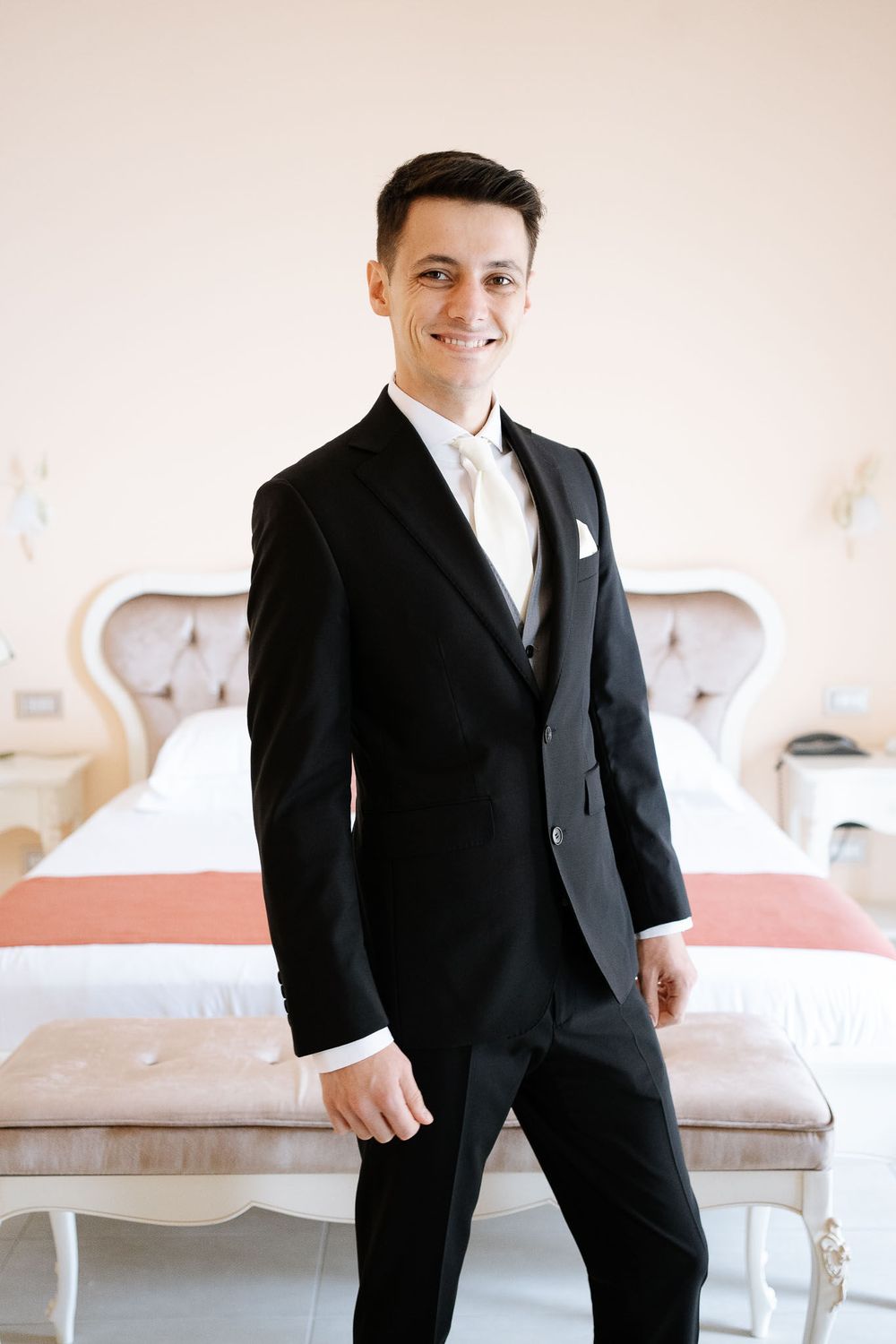 Groom poses confidently in a black tuxedo against an elegant white sofa background.
