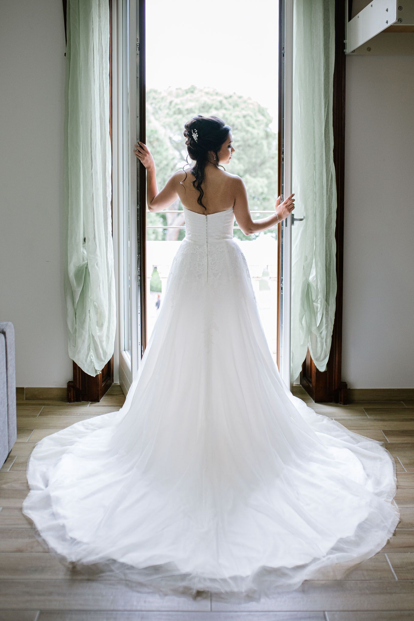 Bride in flowing white wedding gown stands at window, showing dramatic train of her dress.
