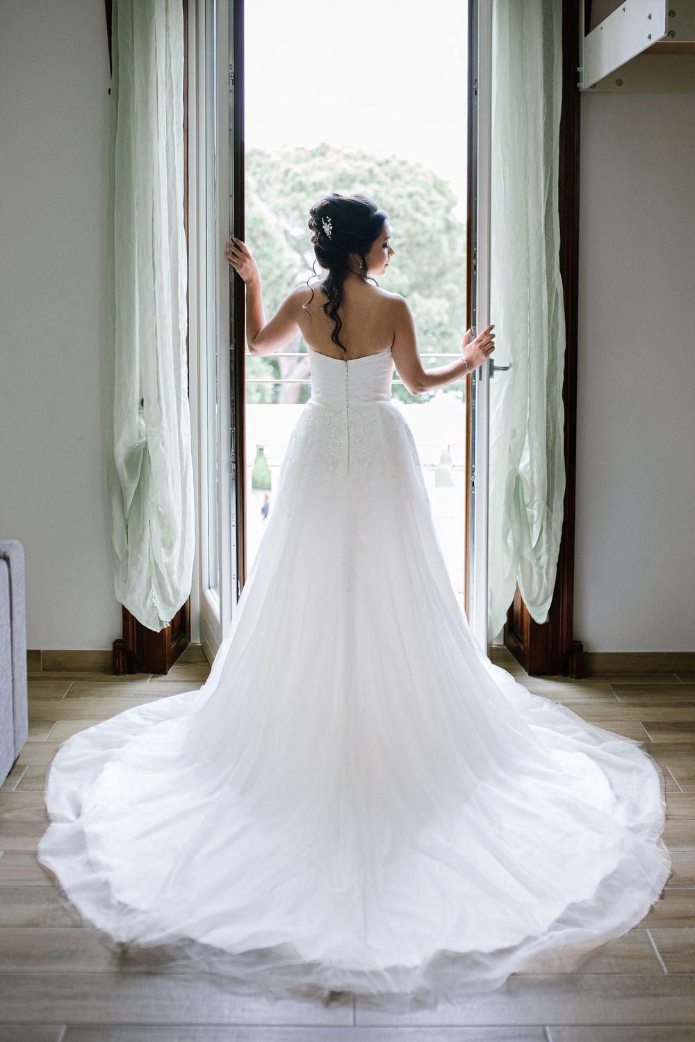 Bride in flowing white wedding gown stands at window, showing dramatic train of her dress.