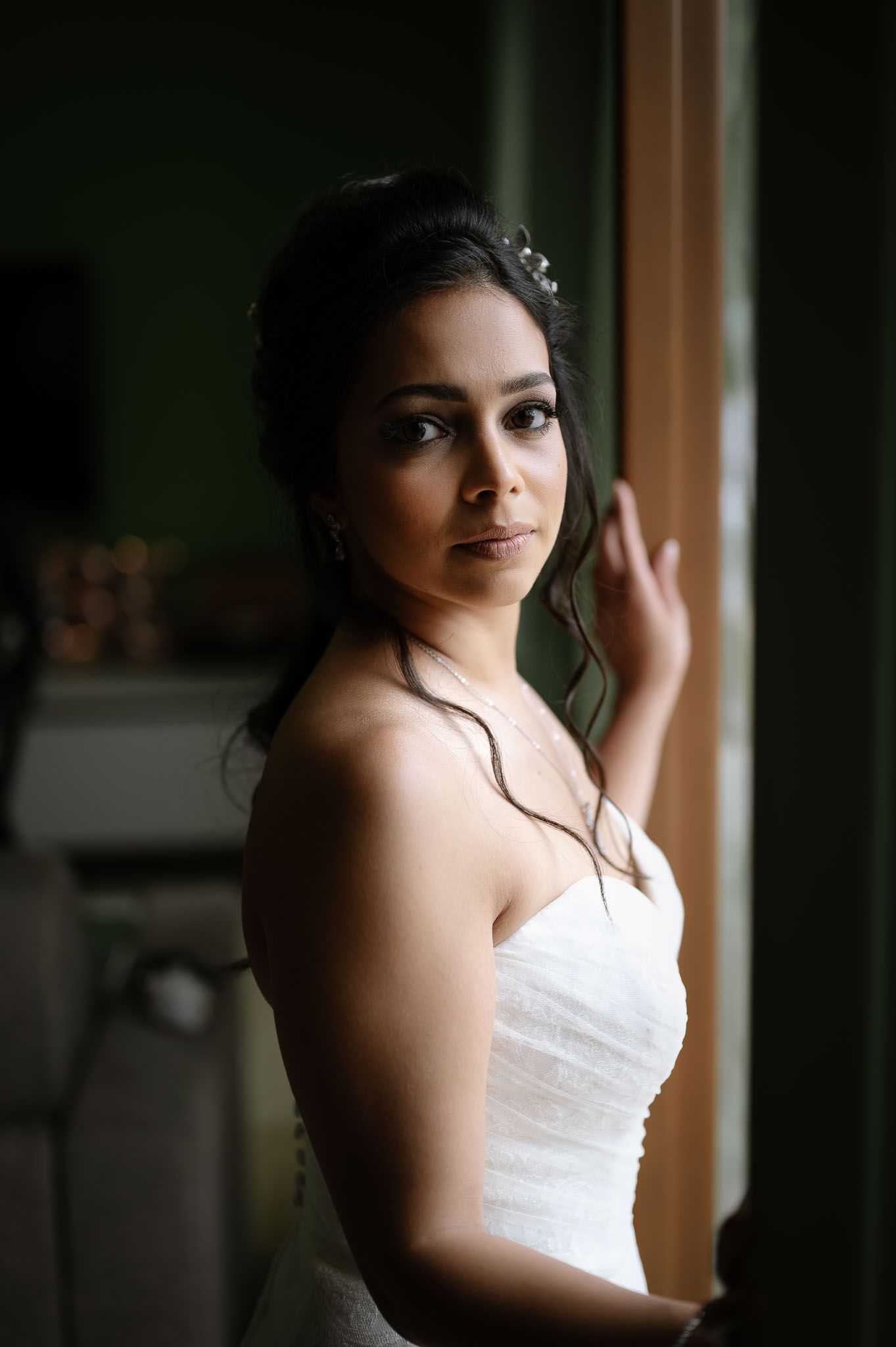 Bride in strapless white gown poses by window with soft natural lighting.