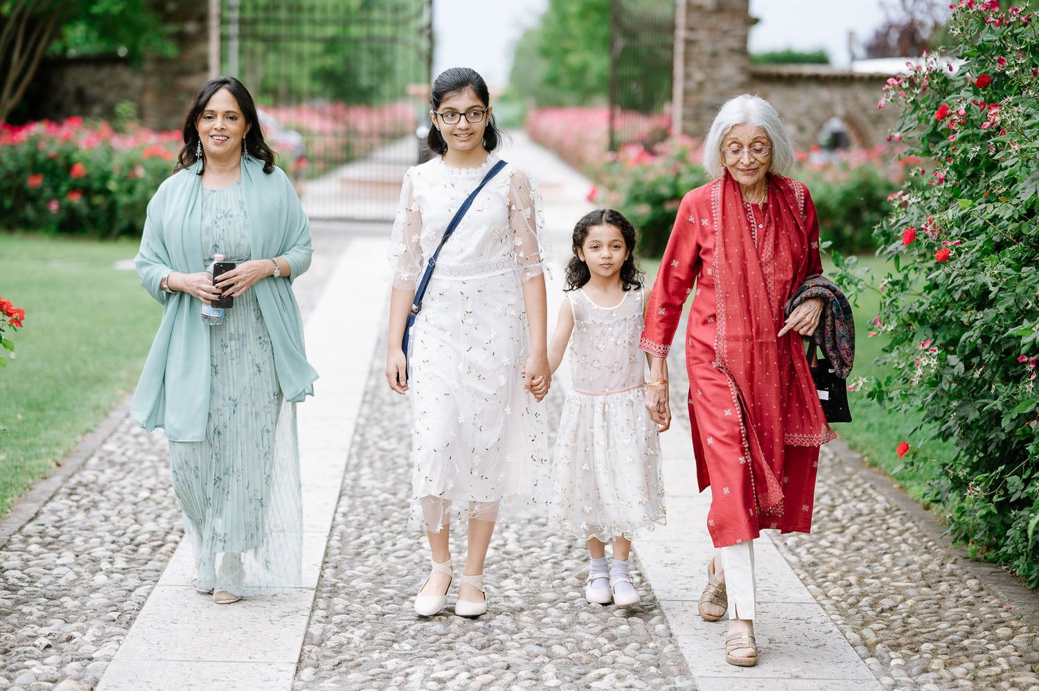 Multi-generational family walking together on garden path in colorful formal attire.