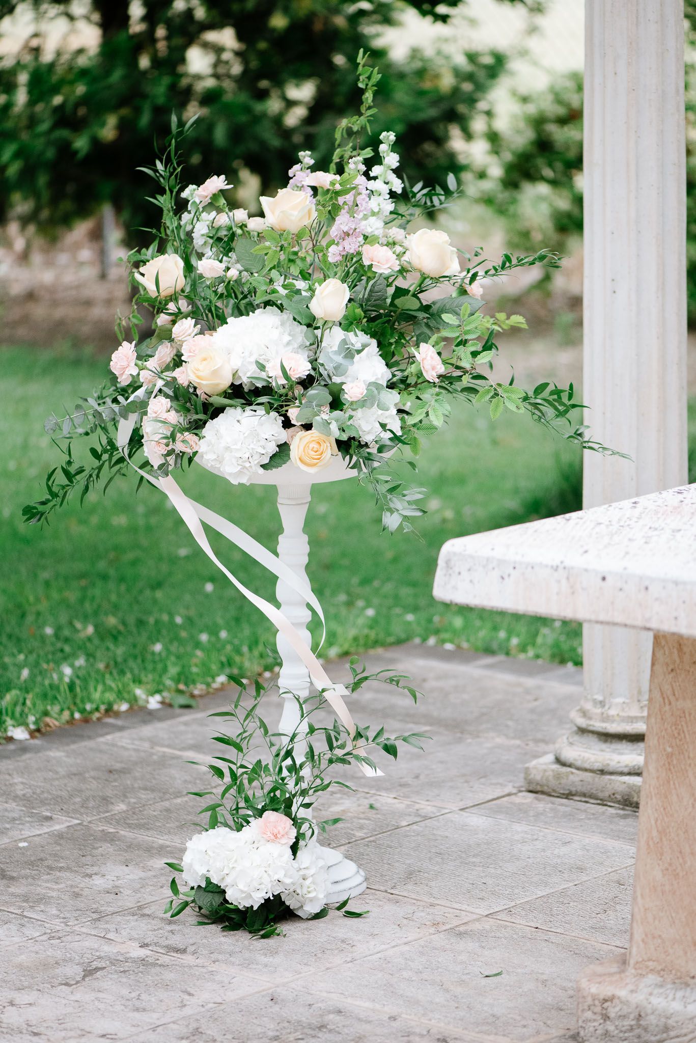 Elegant white floral arrangement with roses and greenery on decorative pedestal.