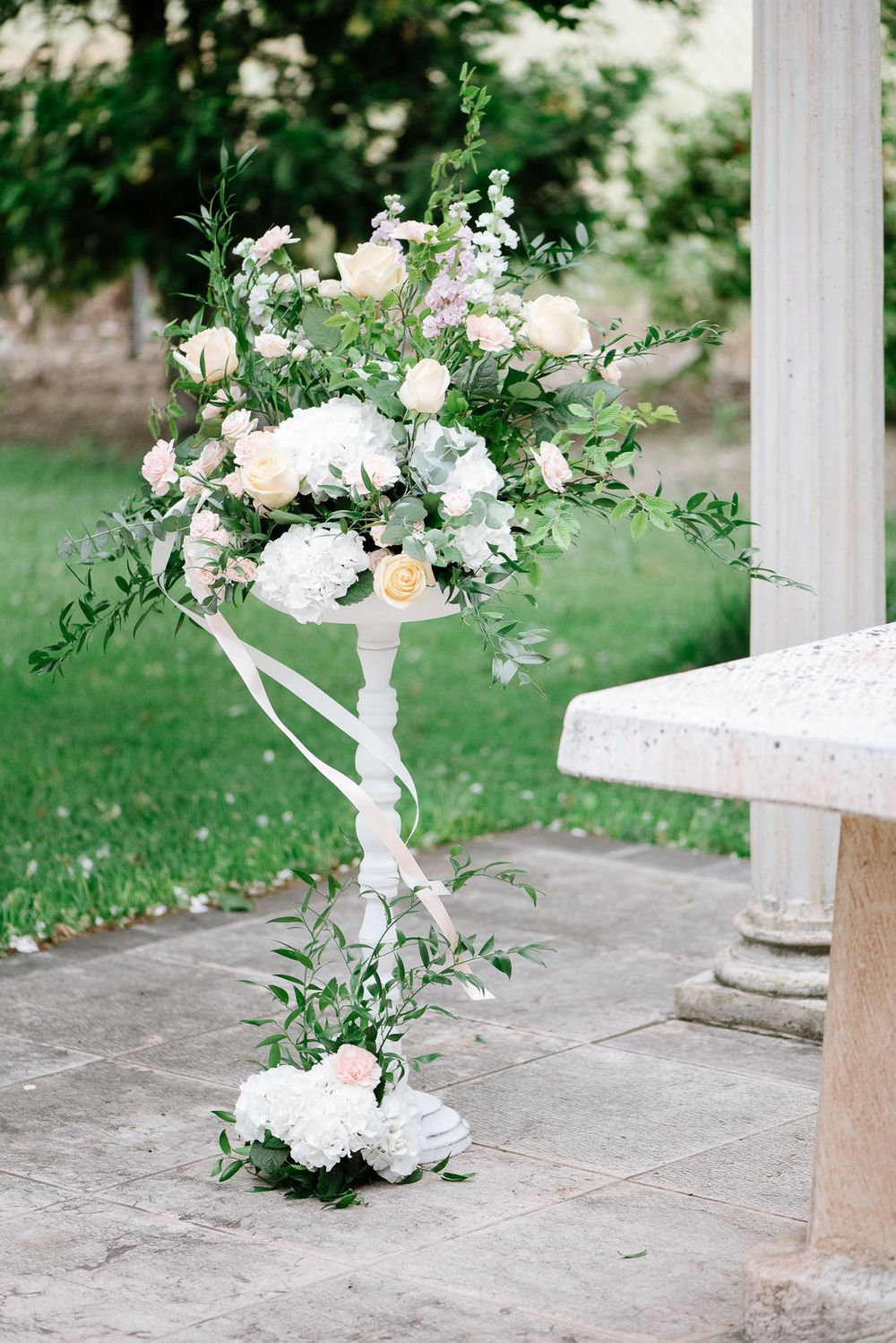 Elegant white floral arrangement with roses and greenery on decorative pedestal.