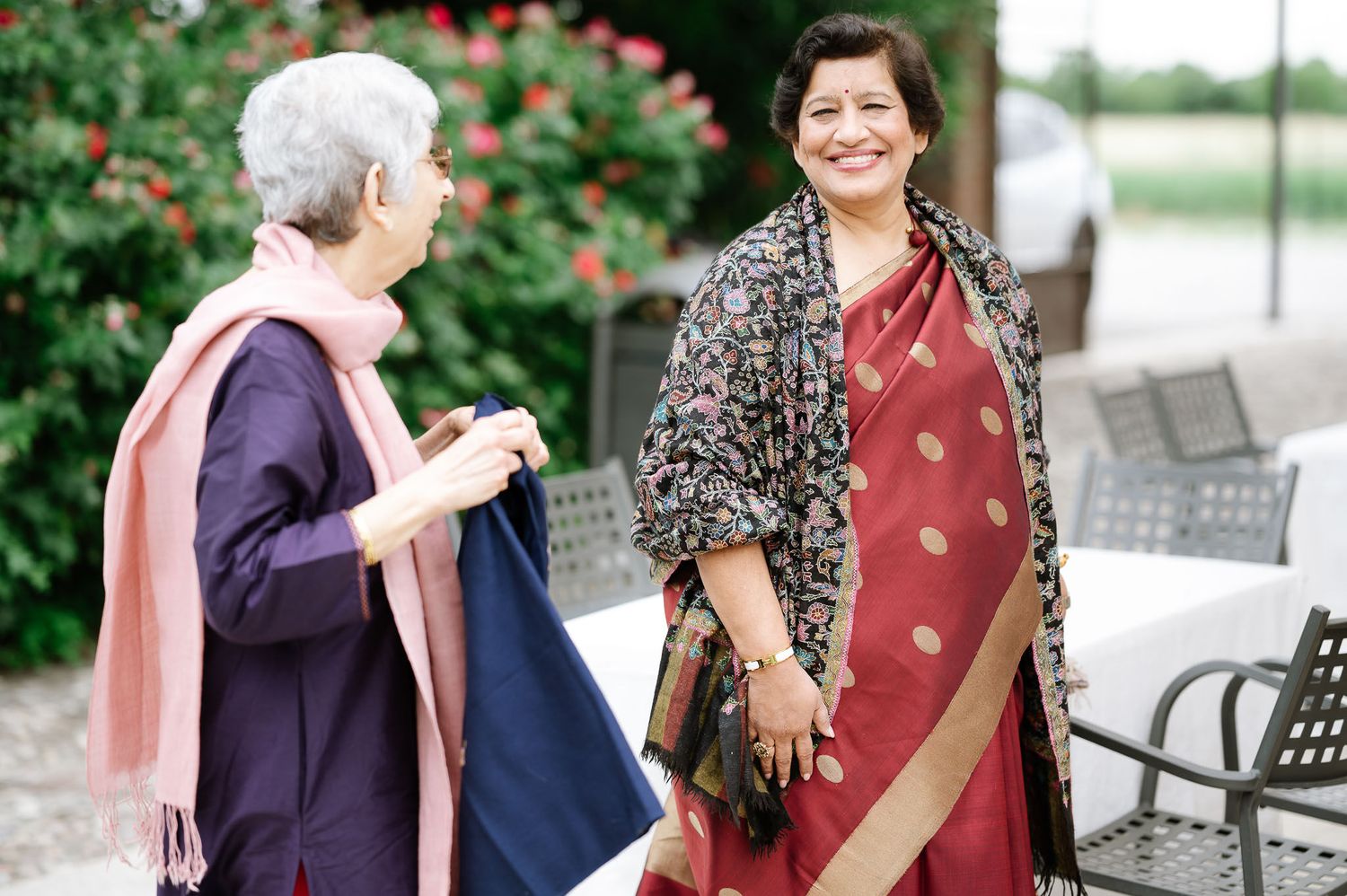 Two women in traditional attire having a conversation outdoors with blooming flowers in the background.