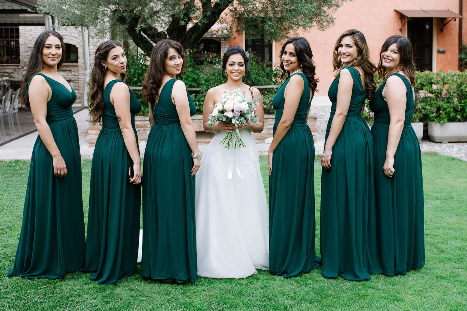 Bridal party in emerald green dresses poses with bride in white on manicured lawn.
