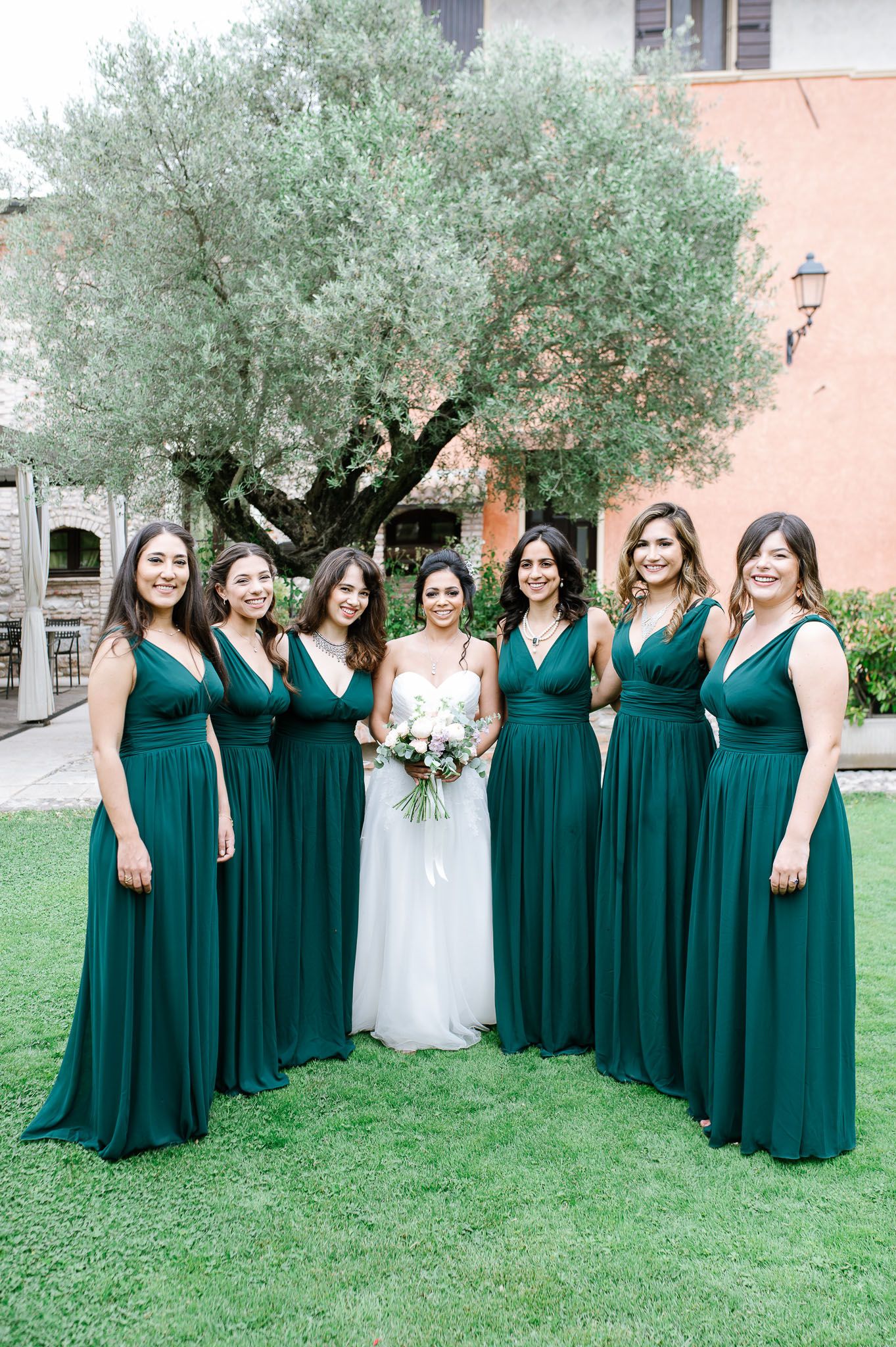 Wedding party in matching emerald gowns gathered around bride under olive tree.
