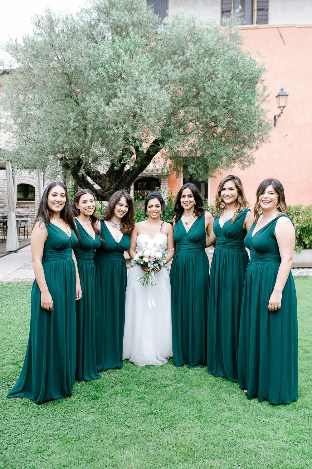 Wedding party in matching emerald gowns gathered around bride under olive tree.