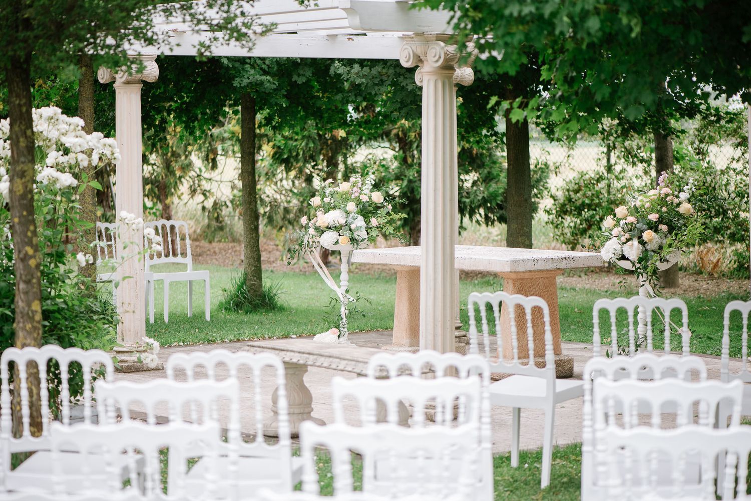 Outdoor wedding ceremony setup with white chairs and classical columns under green trees.
