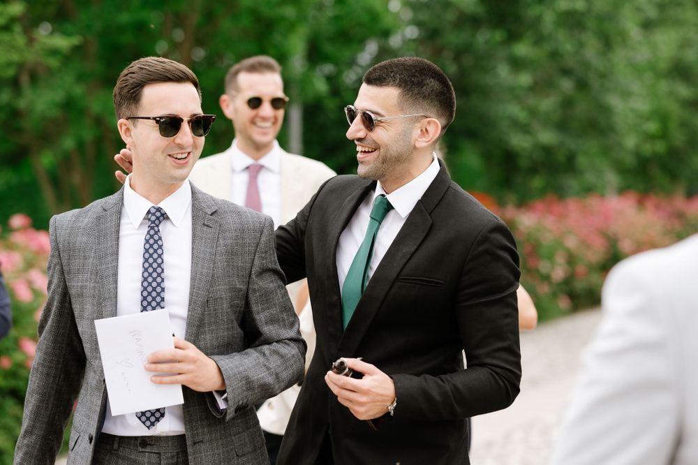 Guests in formal suits sharing a laugh at outdoor wedding event.
