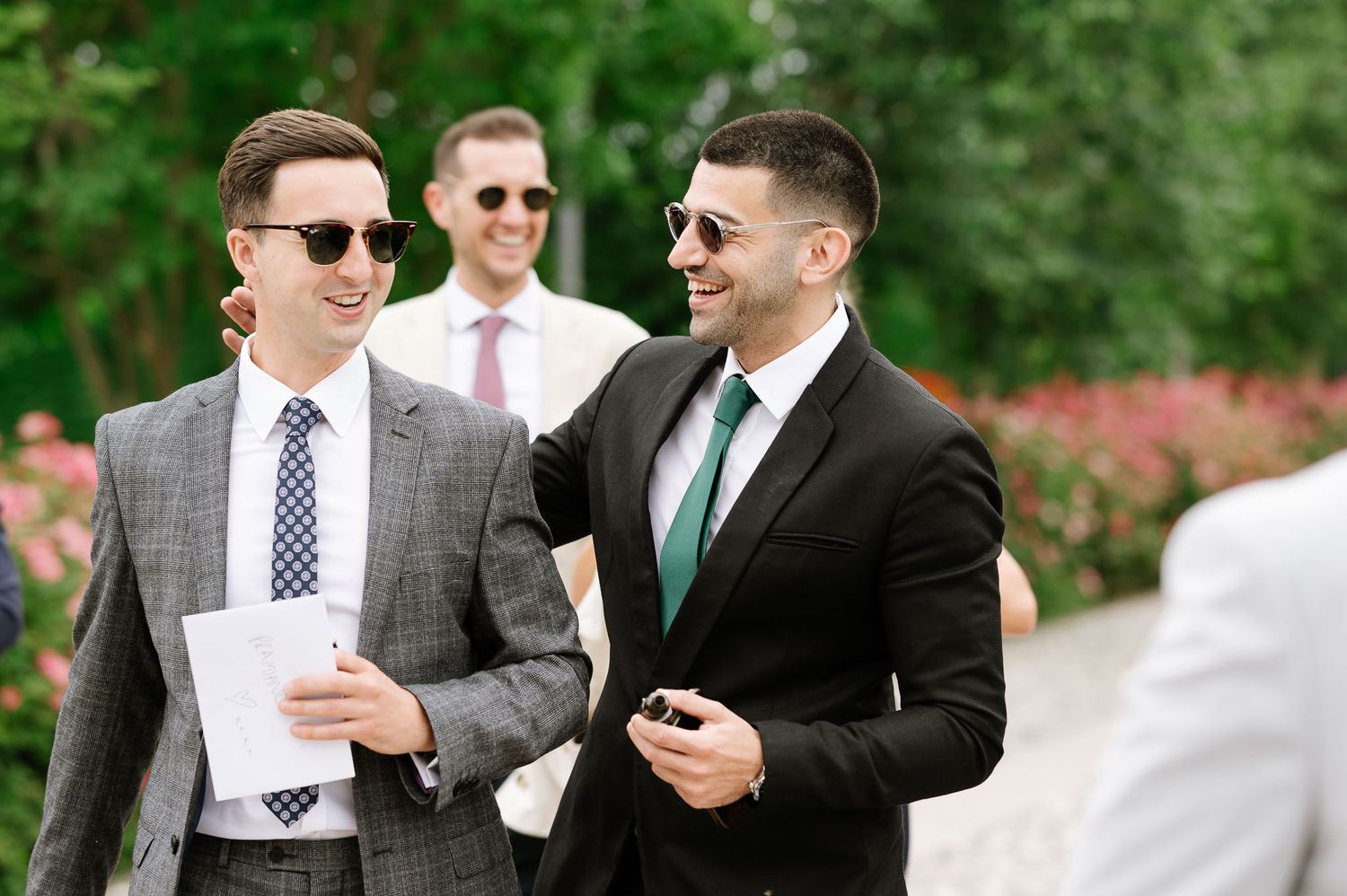 Guests in formal suits sharing a laugh at outdoor wedding event.